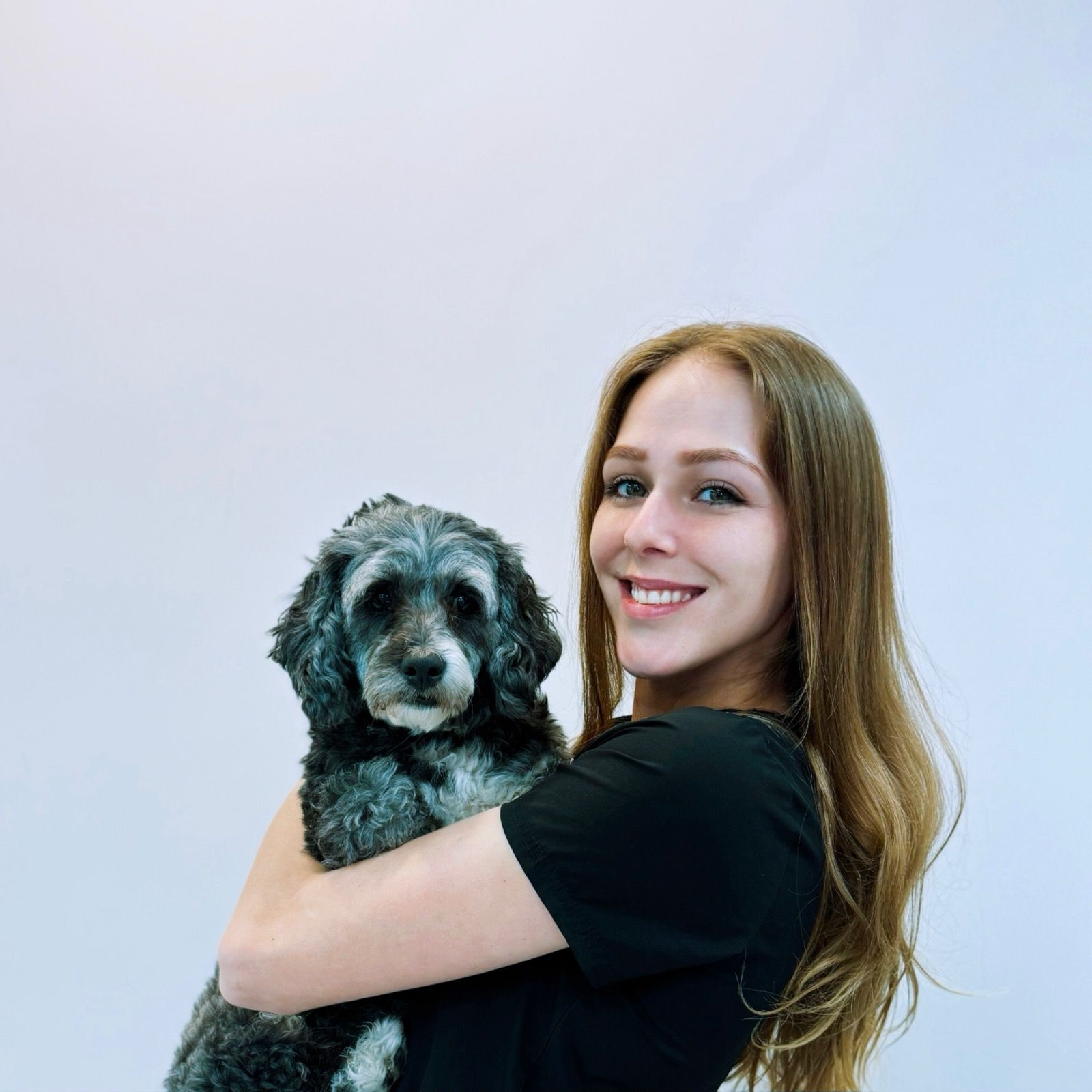 A smiling person with long light brown hair holding a small, curly-haired, grey and black dog against a white background.