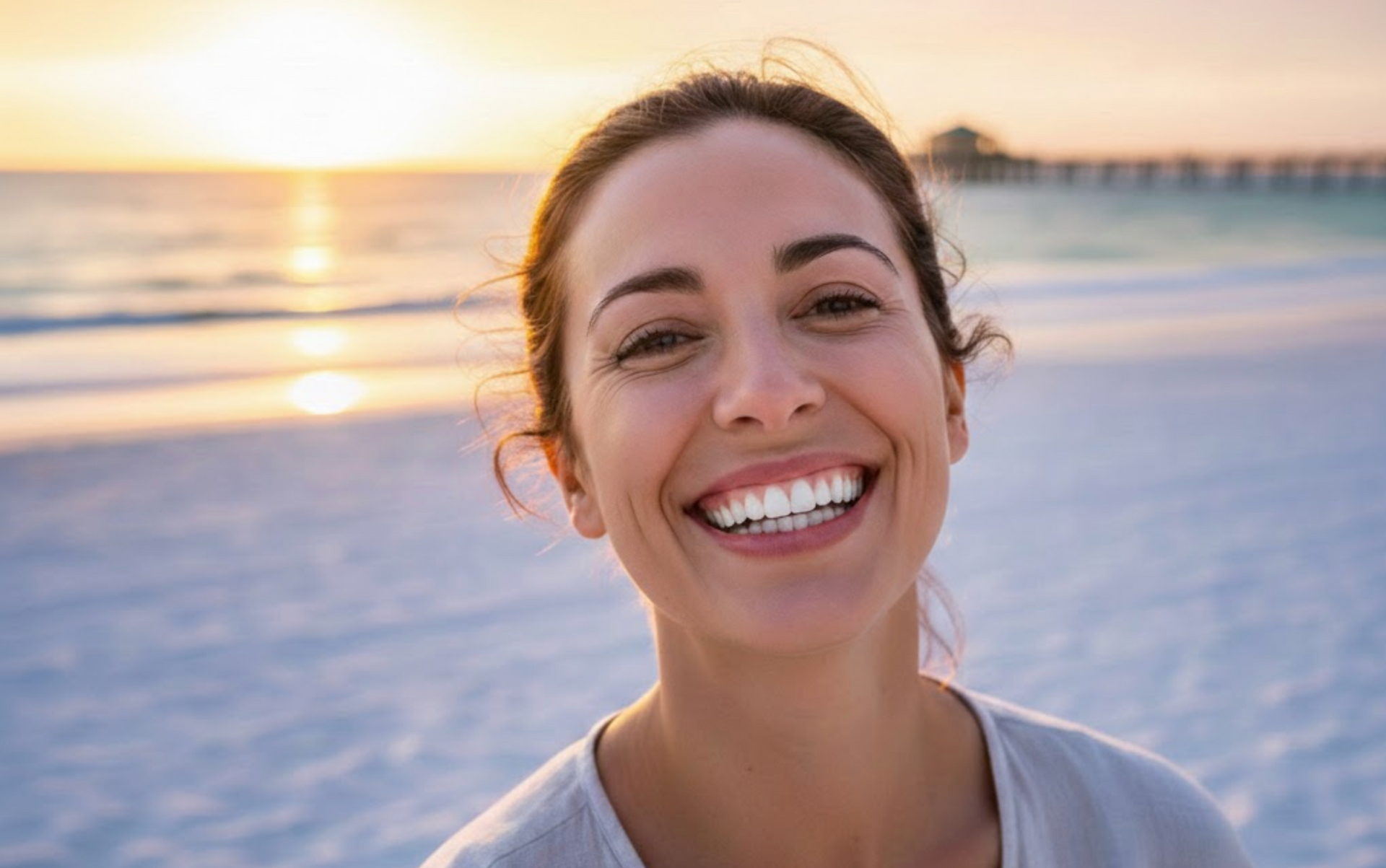 A smiling person stands on a sandy beach at sunset, with the ocean and a pier visible in the background.
