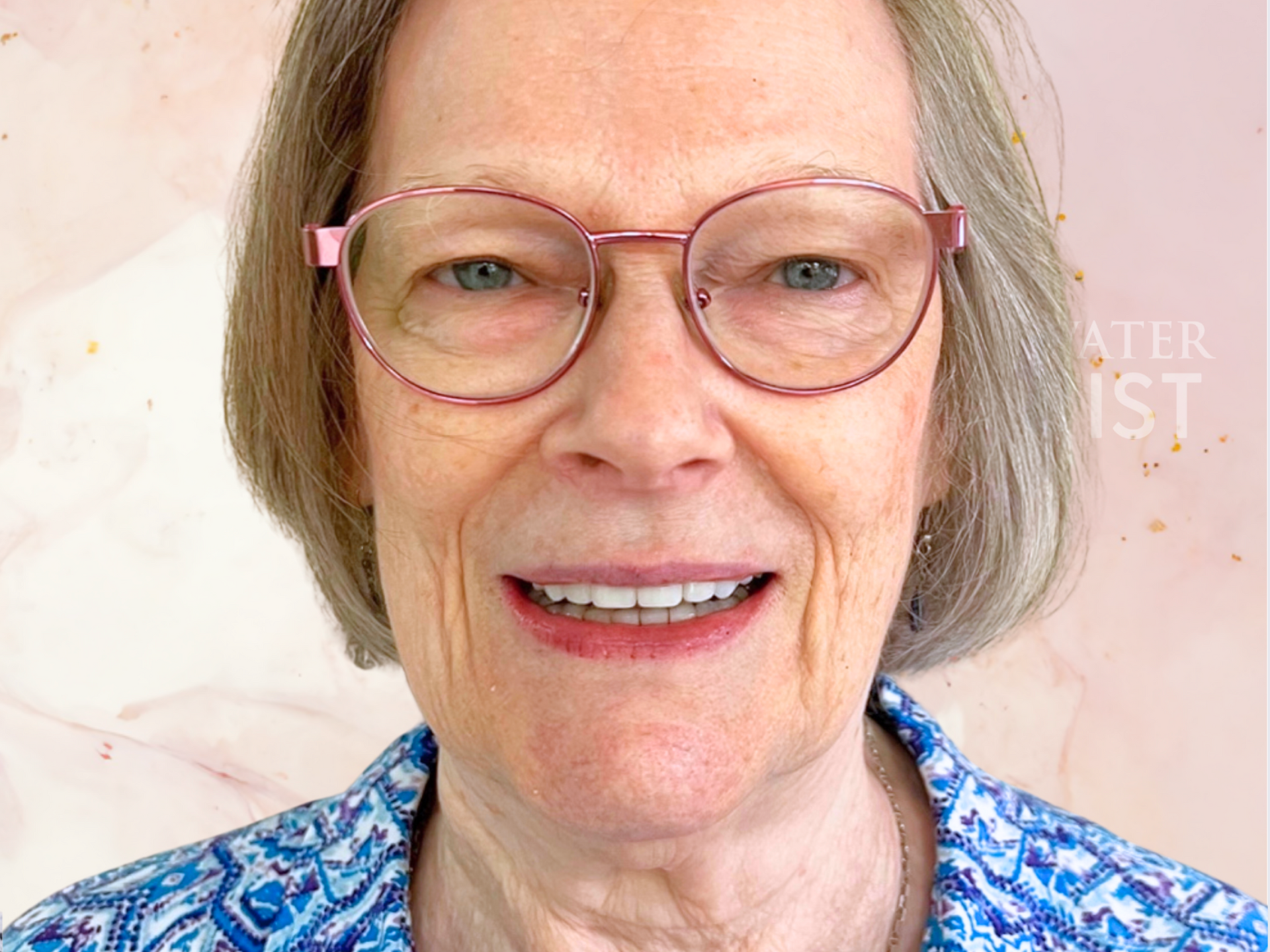 A smiling person wearing glasses and a blue patterned top against a soft pink background.