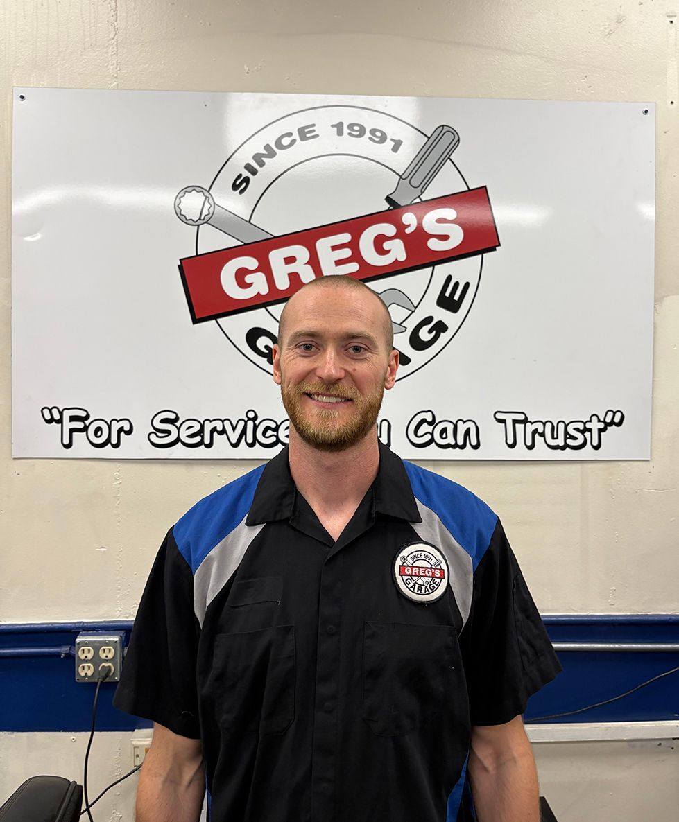 Man in mechanic shop, wearing black and blue shirt, smiles at camera while working at a computer. | Greg's Garage
