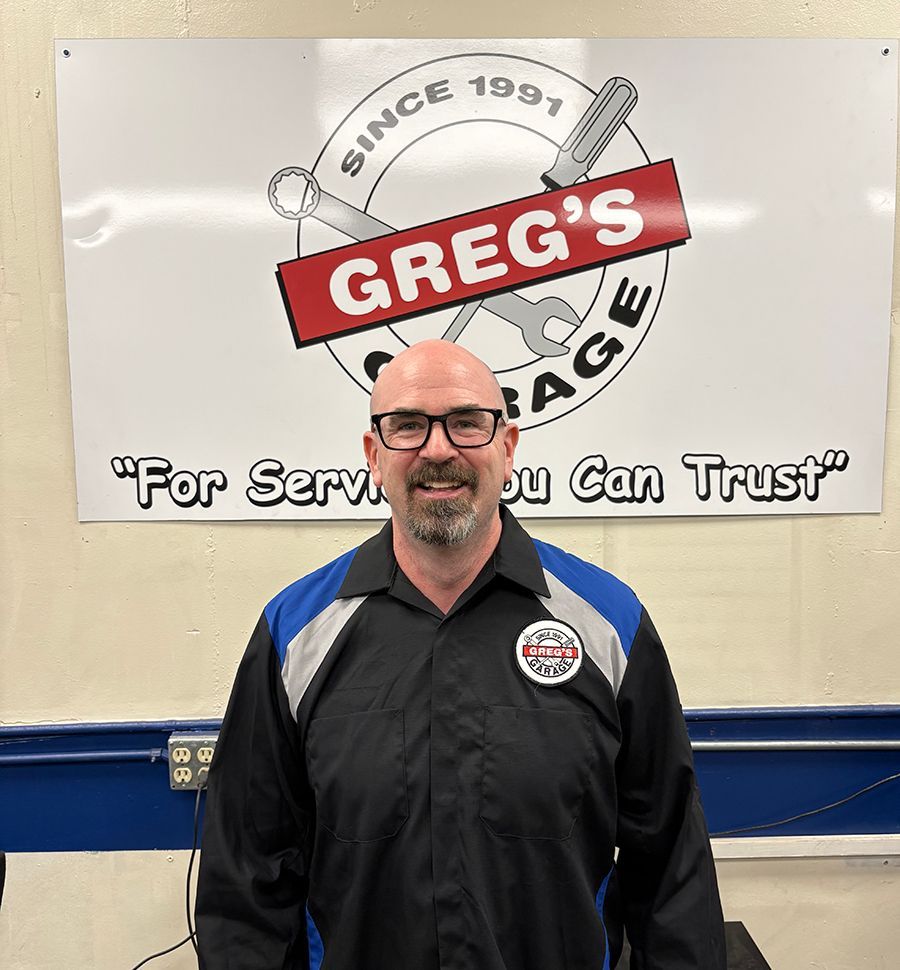 Man in mechanic shop, wearing black and blue shirt, smiles at camera while working at a computer. | Greg's Garage
