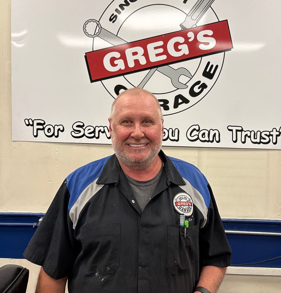 Man in mechanic shop, wearing black and blue shirt, smiles at camera while working at a computer. | Greg's Garage