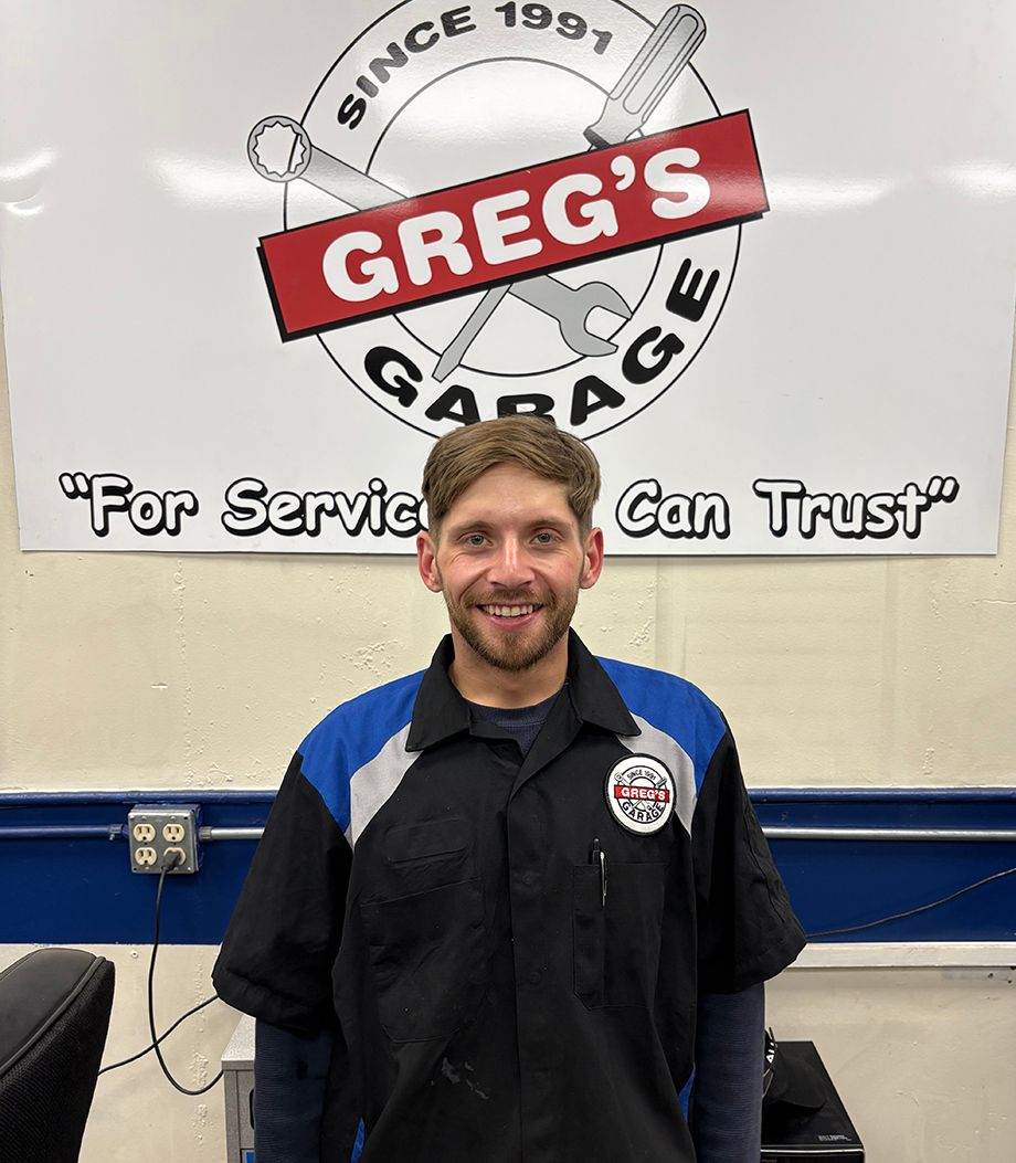 Man in mechanic shop, wearing black and blue shirt, smiles at camera while working at a computer. | Greg's Garage