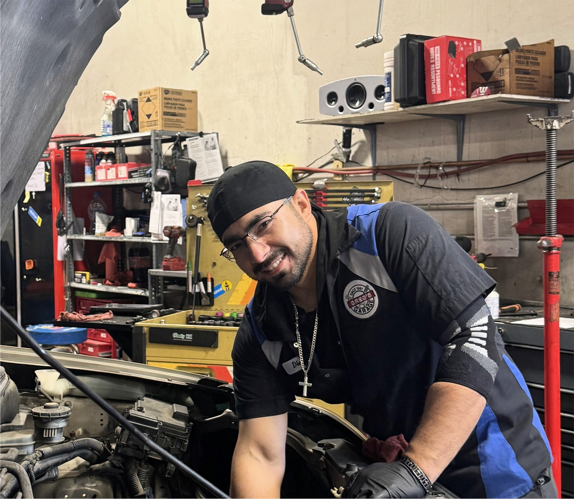 Mechanic smiling while working on a car engine in a garage, wearing a black hat and blue/black uniform. | Greg's Garage