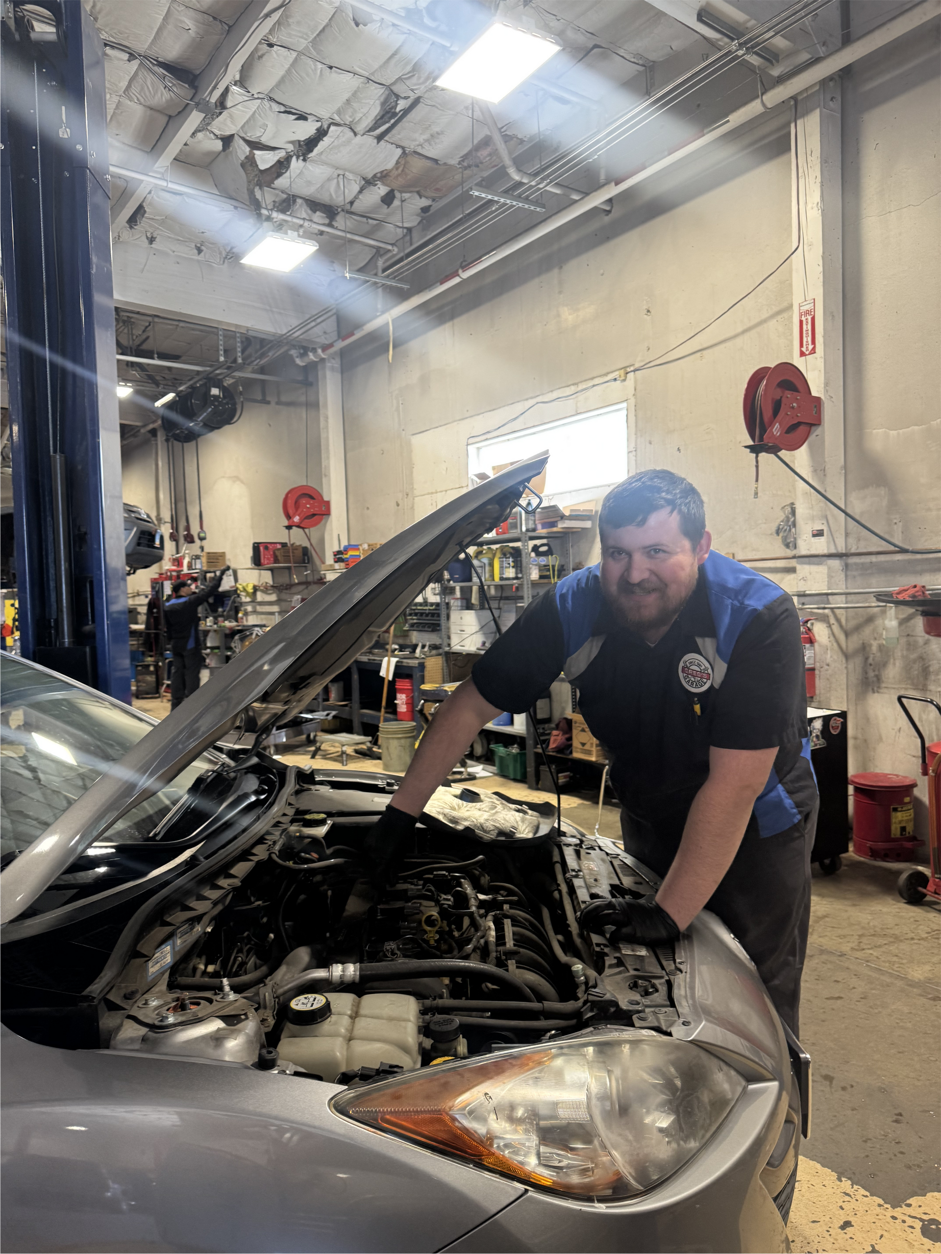 Mechanic in blue shirt smiles while working on a car engine inside a garage. | Greg's Garage