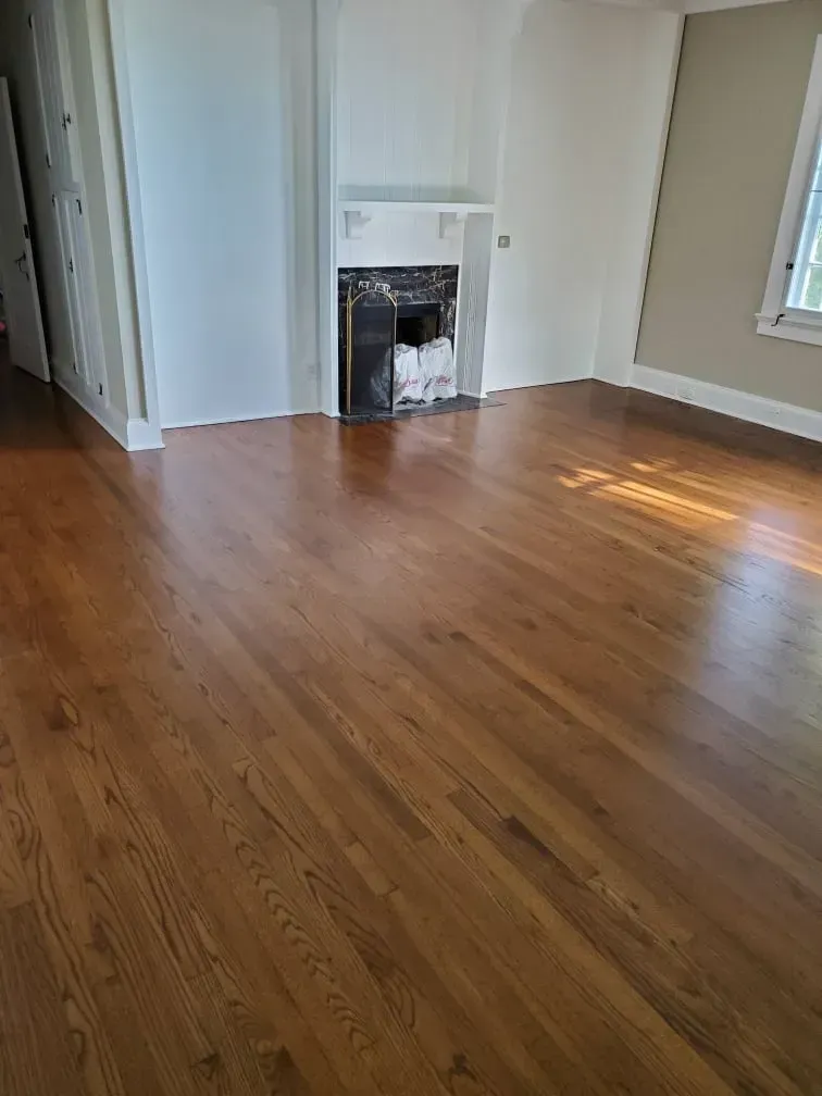 Empty living room with hardwood floors, white walls, and a small fireplace under a window.