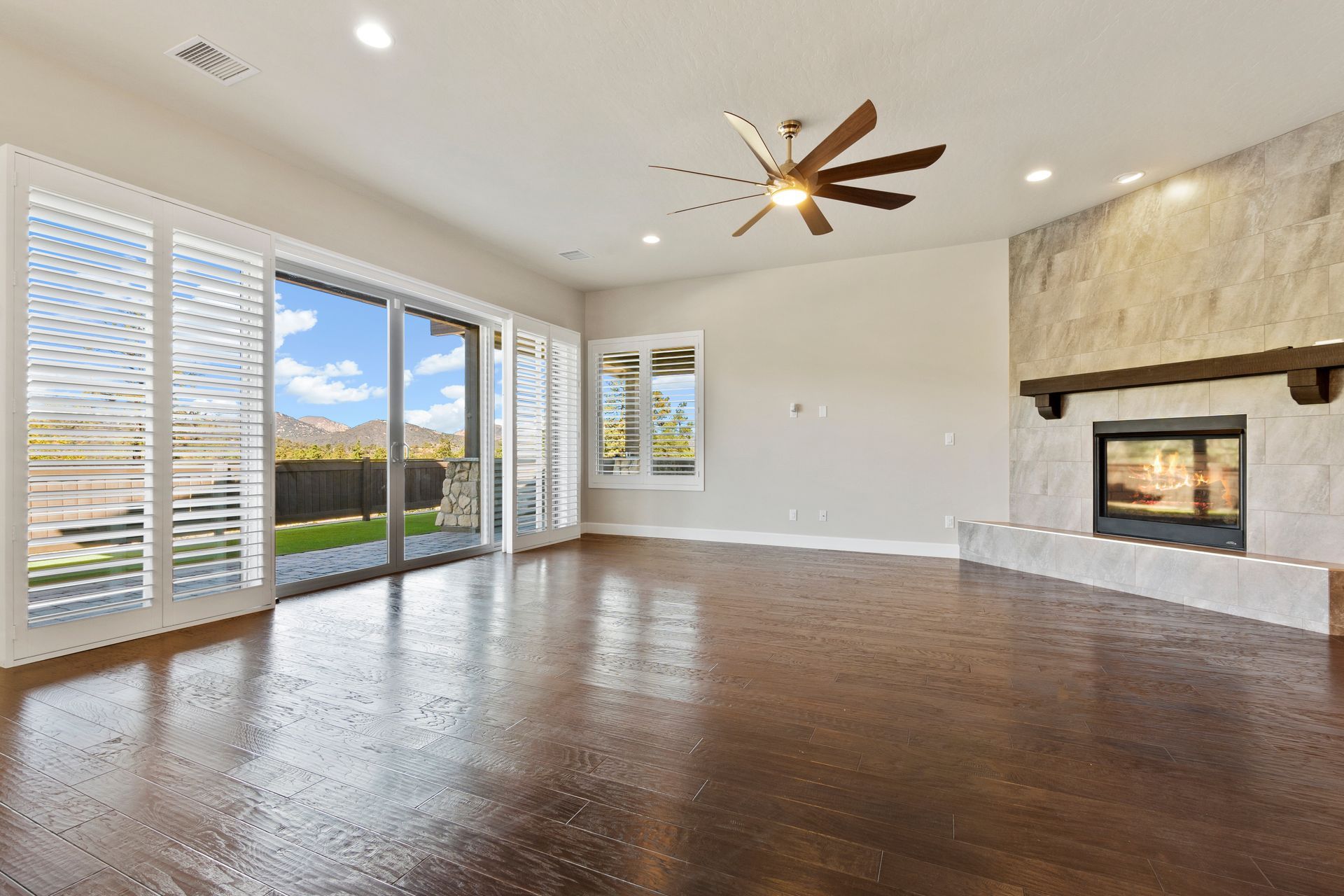 Empty living room with dark wood floors, large windows, ceiling fan, and stone fireplace
