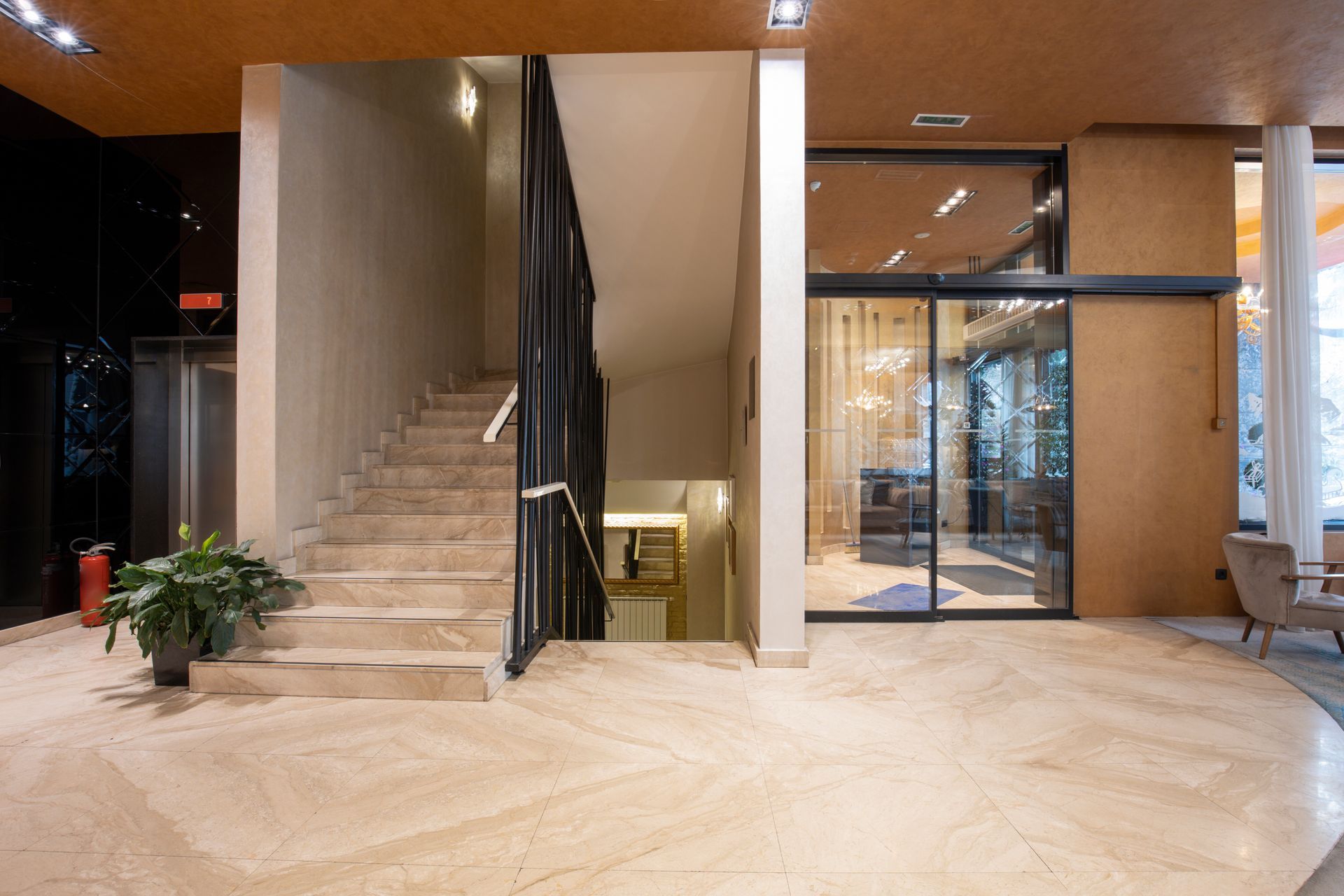 Modern lobby with staircase, black railing, glass doors, and potted plant on a light floor.