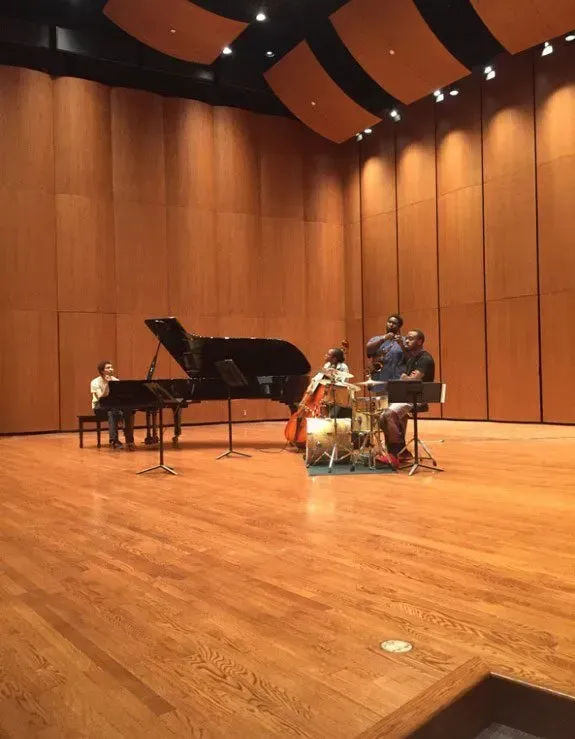 Jazz trio performing on a wooden concert stage with a grand piano and drum set under warm lights.