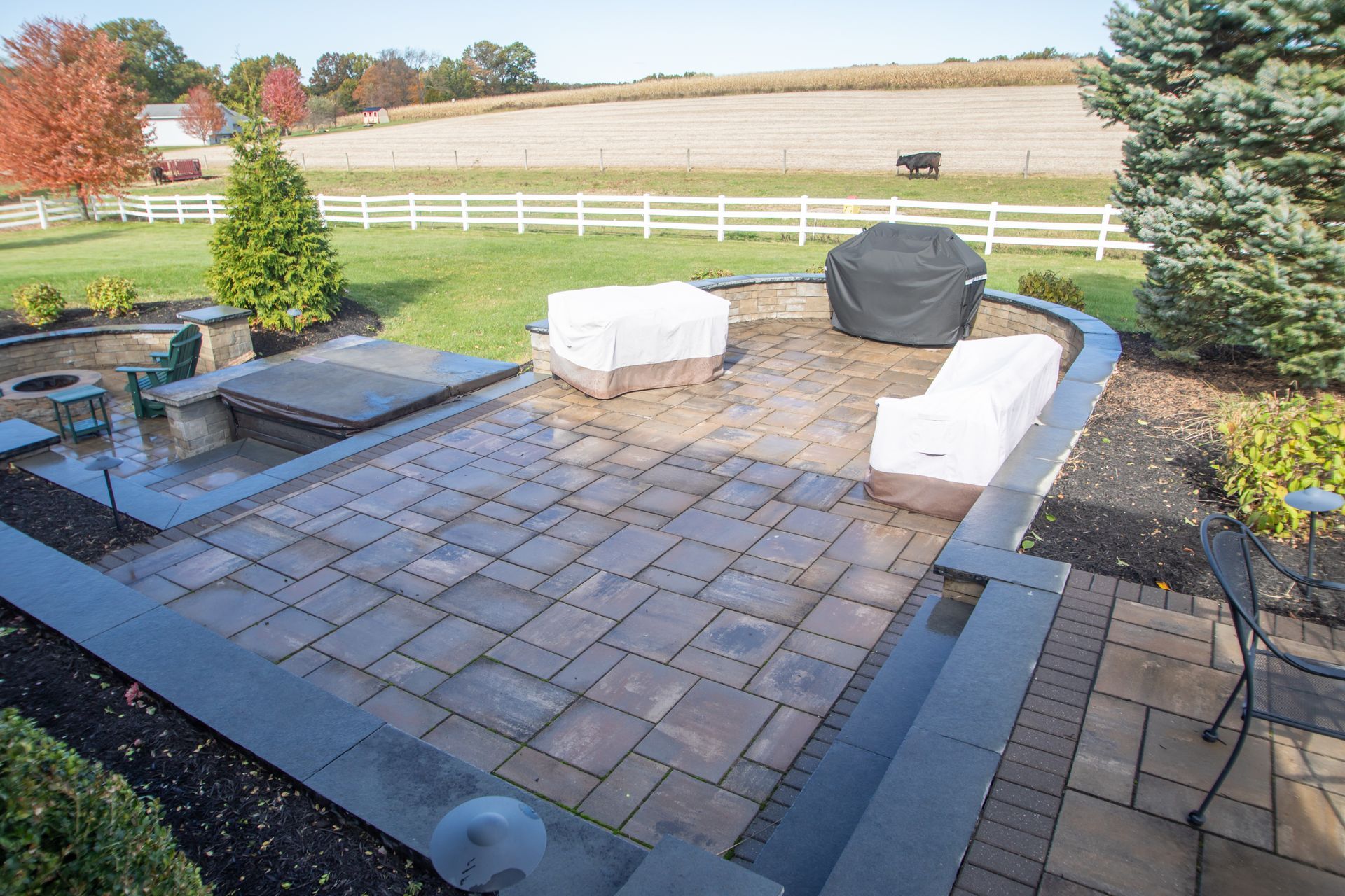 Stone patio with covered furniture, fire pit, and view of a field behind a white fence.