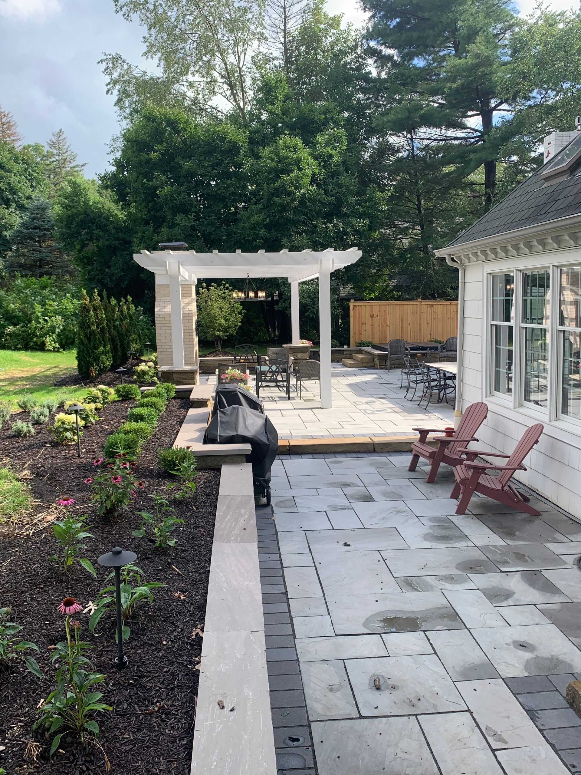 Stone patio with pergola, grill, and Adirondack chairs near a white house and garden.