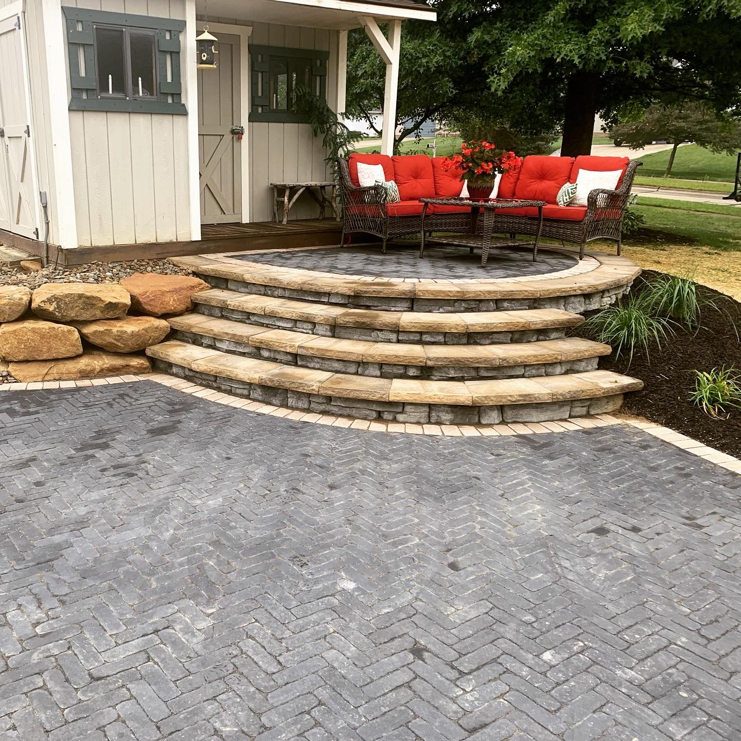 A paved patio area with steps leading to a seating area with red cushions and a small structure.