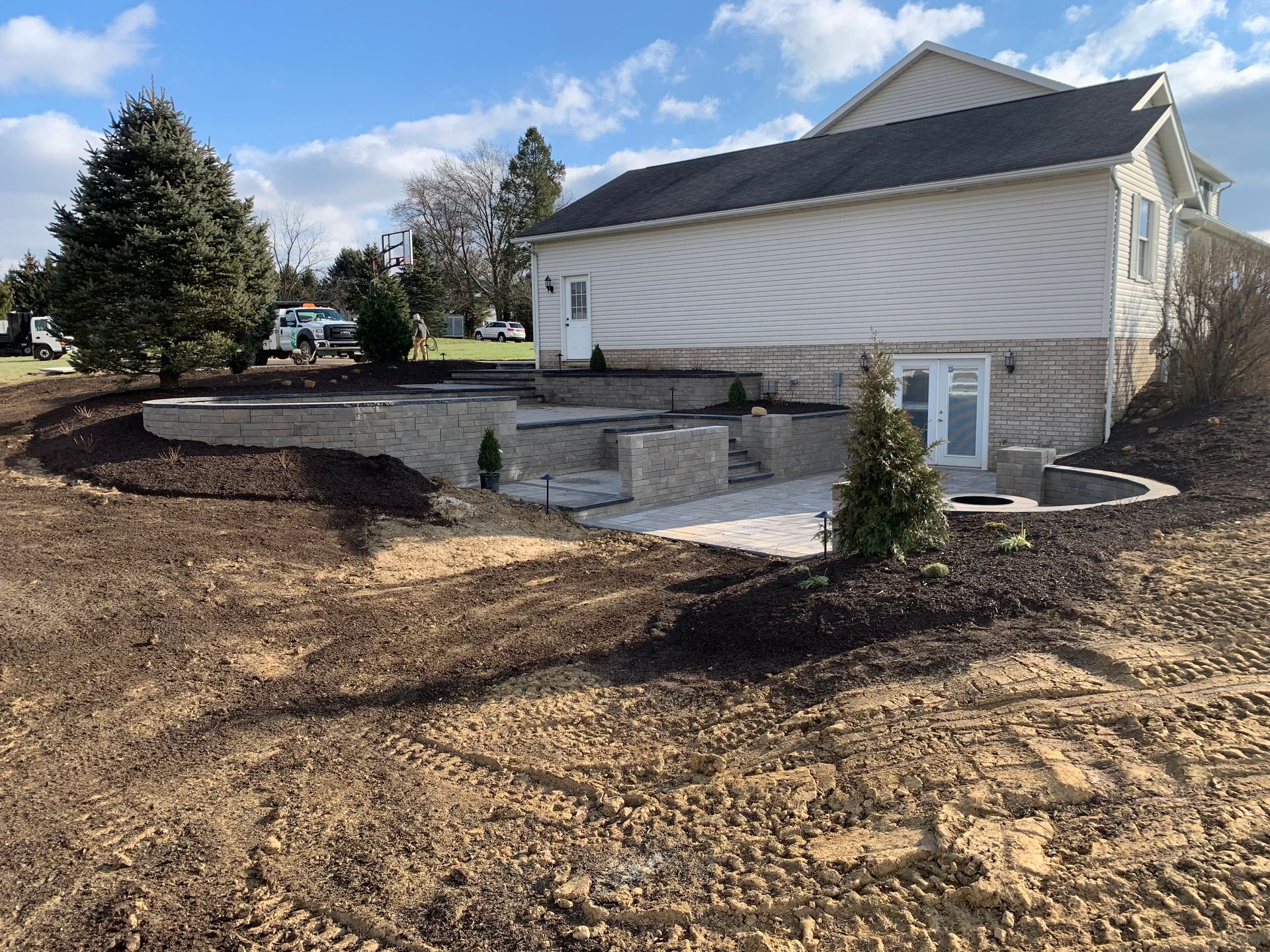 Backyard landscape with retaining walls and patio next to a house with a door and basement windows.