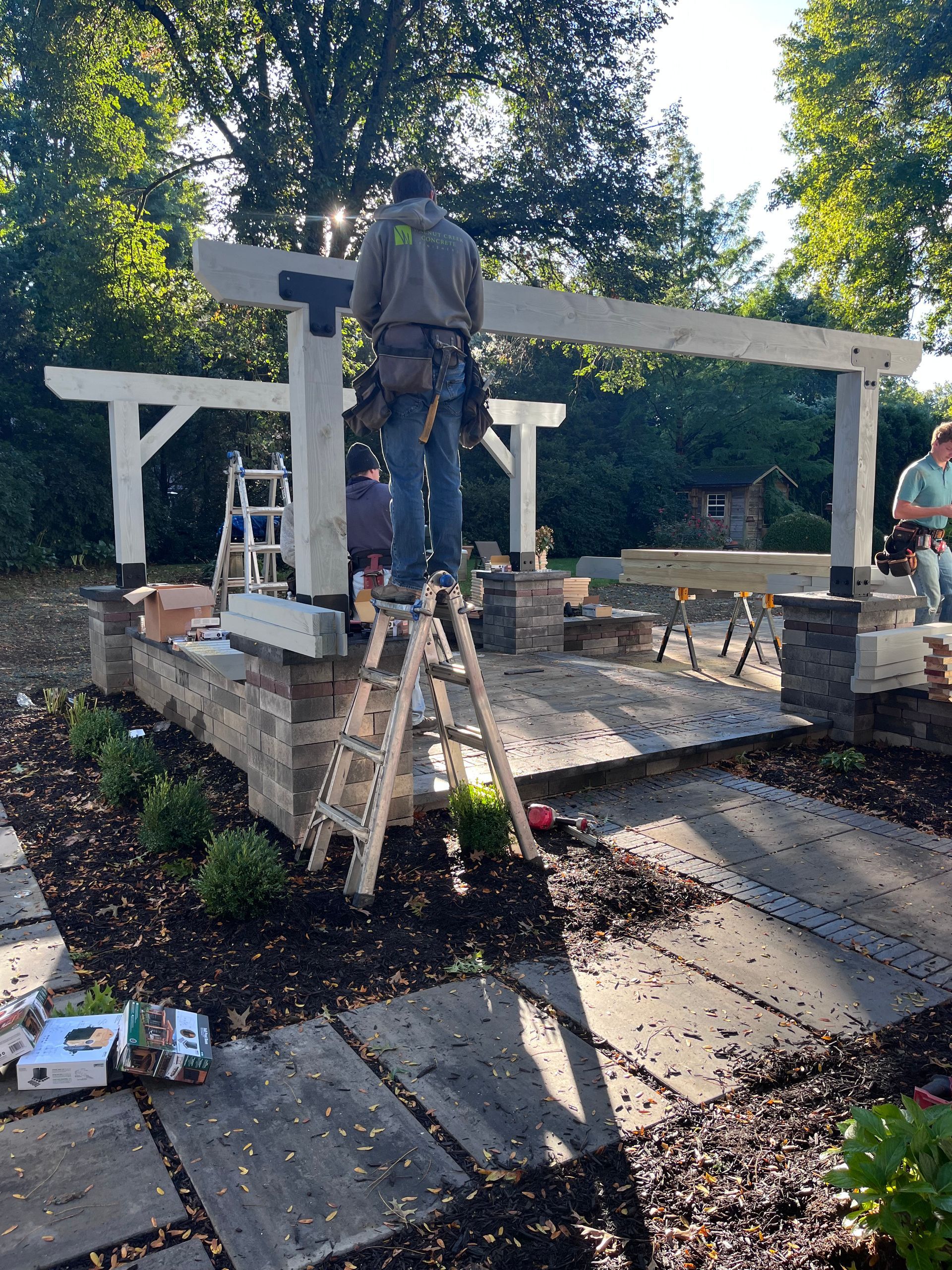 Construction worker on ladder, building a pergola in a yard. Gray stone patio, wood beams, and tools are visible.