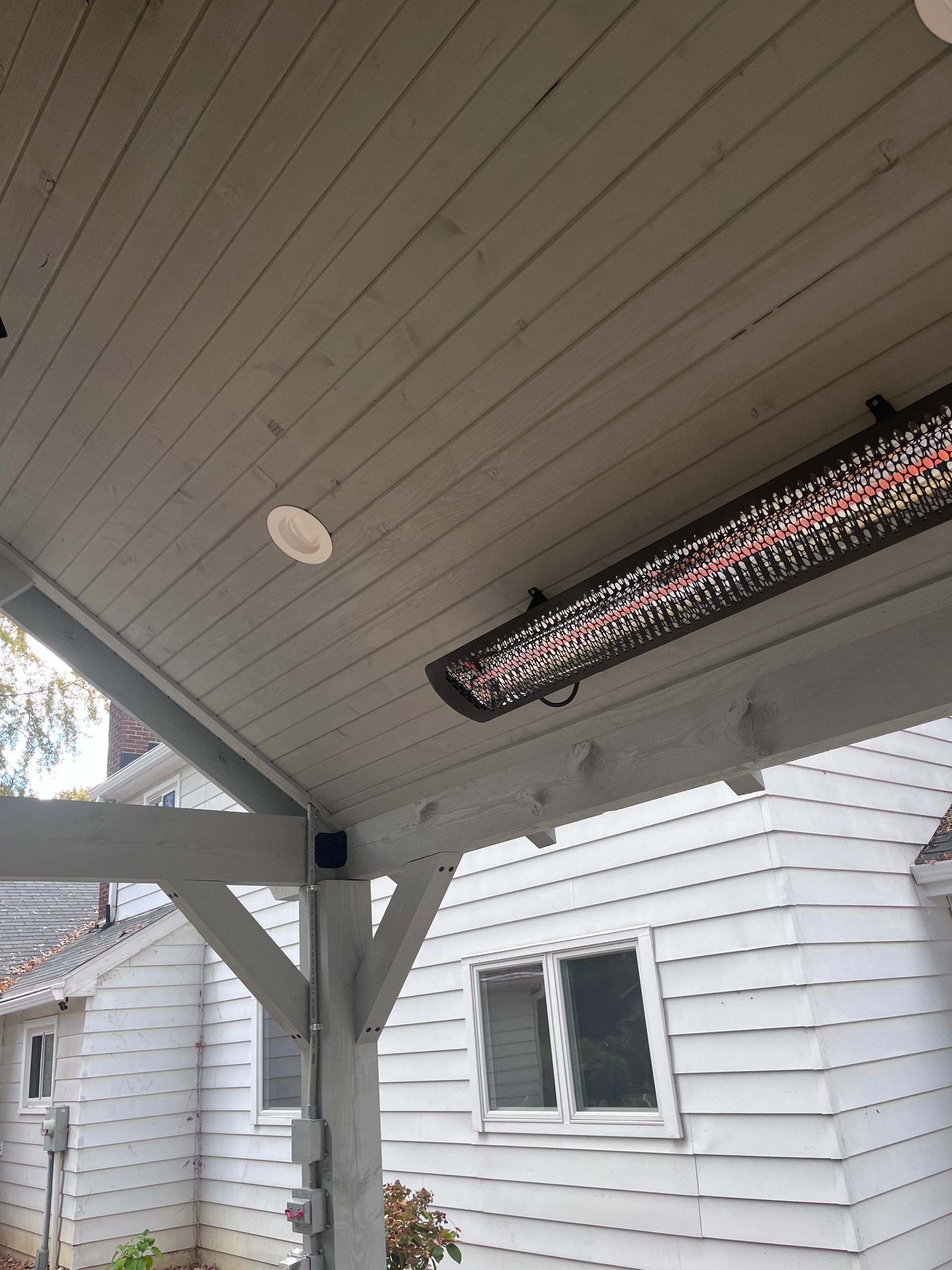 Patio heater mounted on a white, wood-paneled ceiling with a light fixture, near a white house.