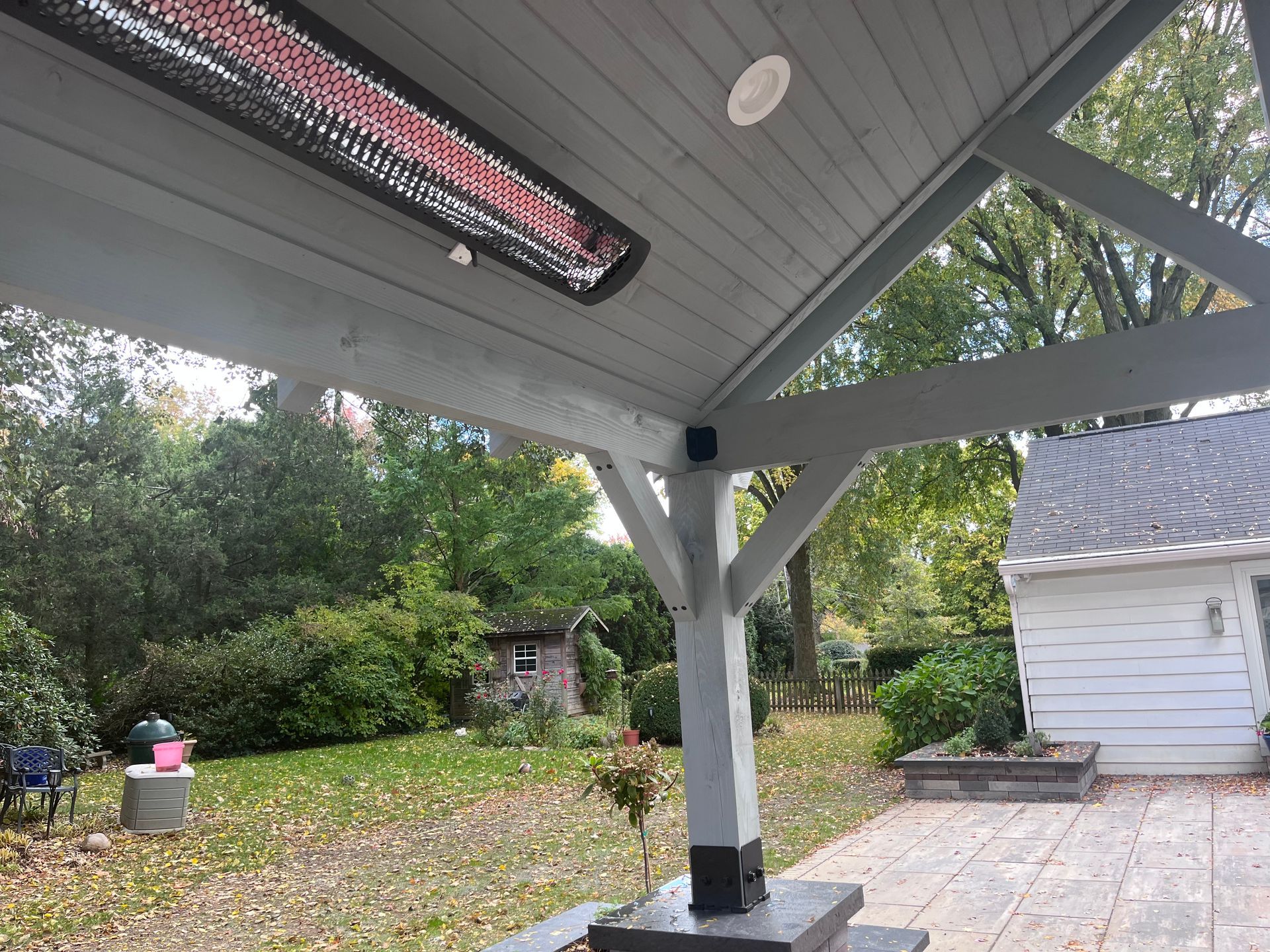 Covered patio with heater, gray beams, backyard view with trees, and stone patio.