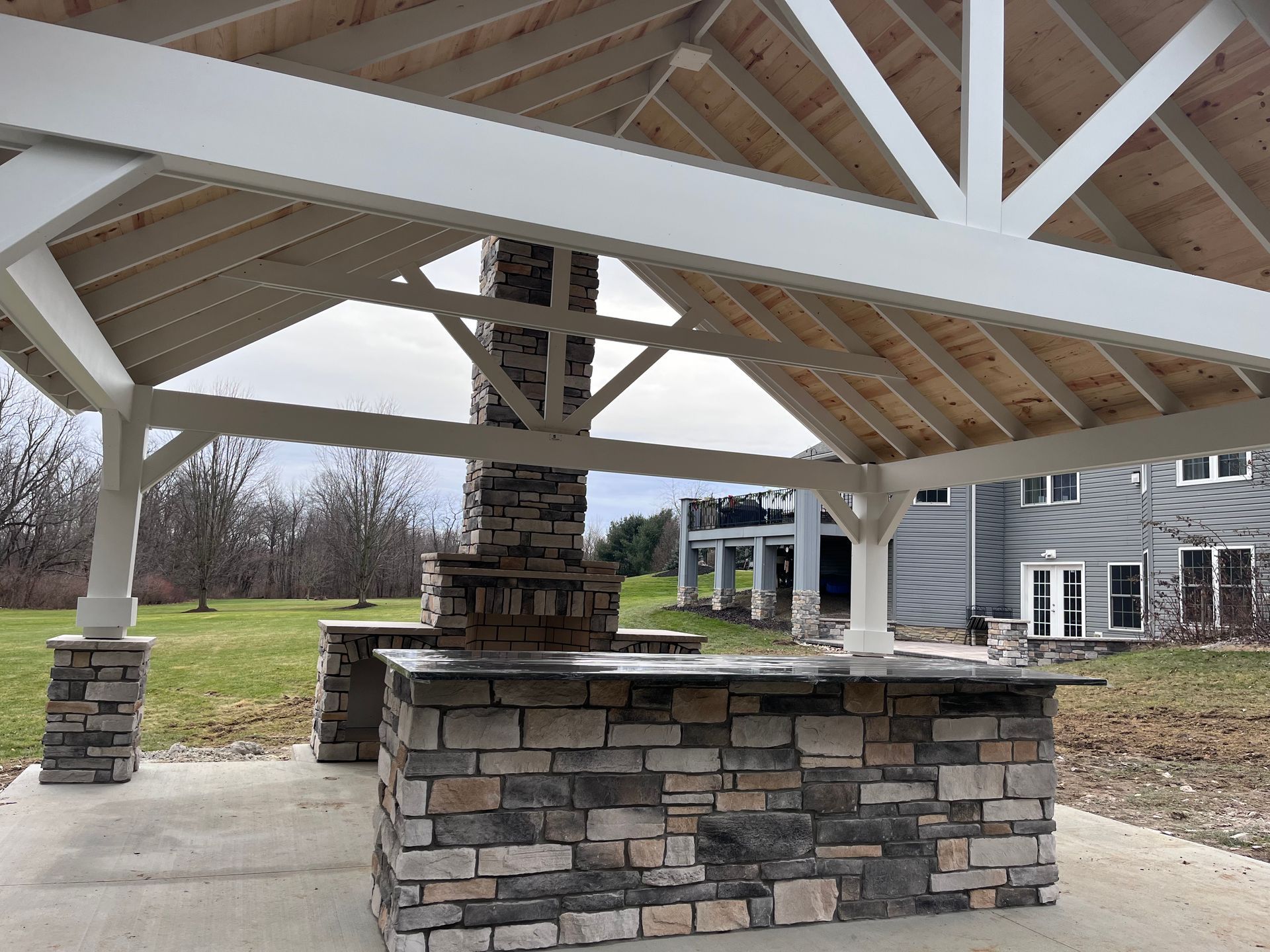 Outdoor stone bar with fireplace under a white-framed wooden pergola with a view of a grassy field and house.