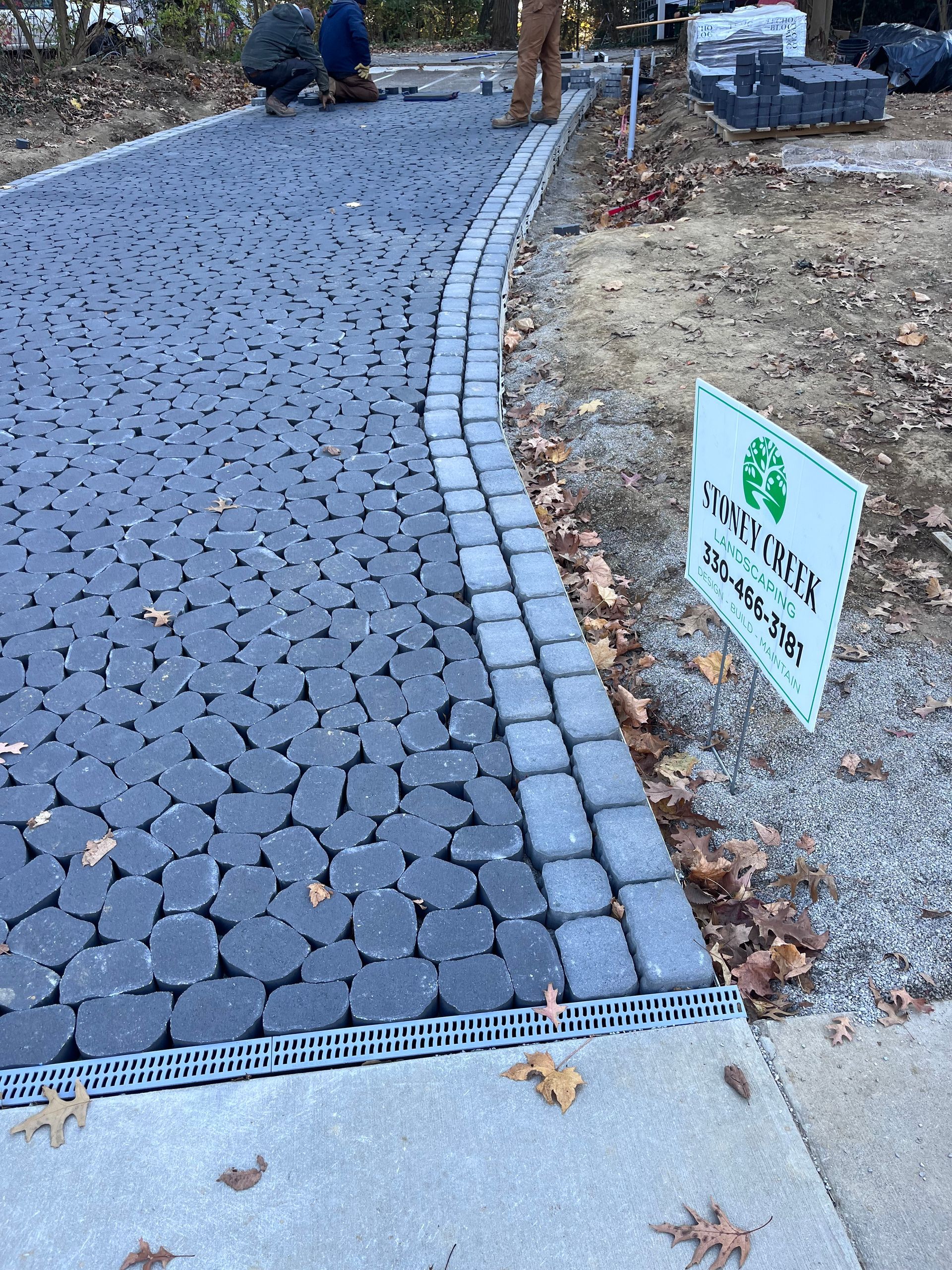 Driveway with permeable paving, edged with pavers, bordered by a concrete curb; workers in background.