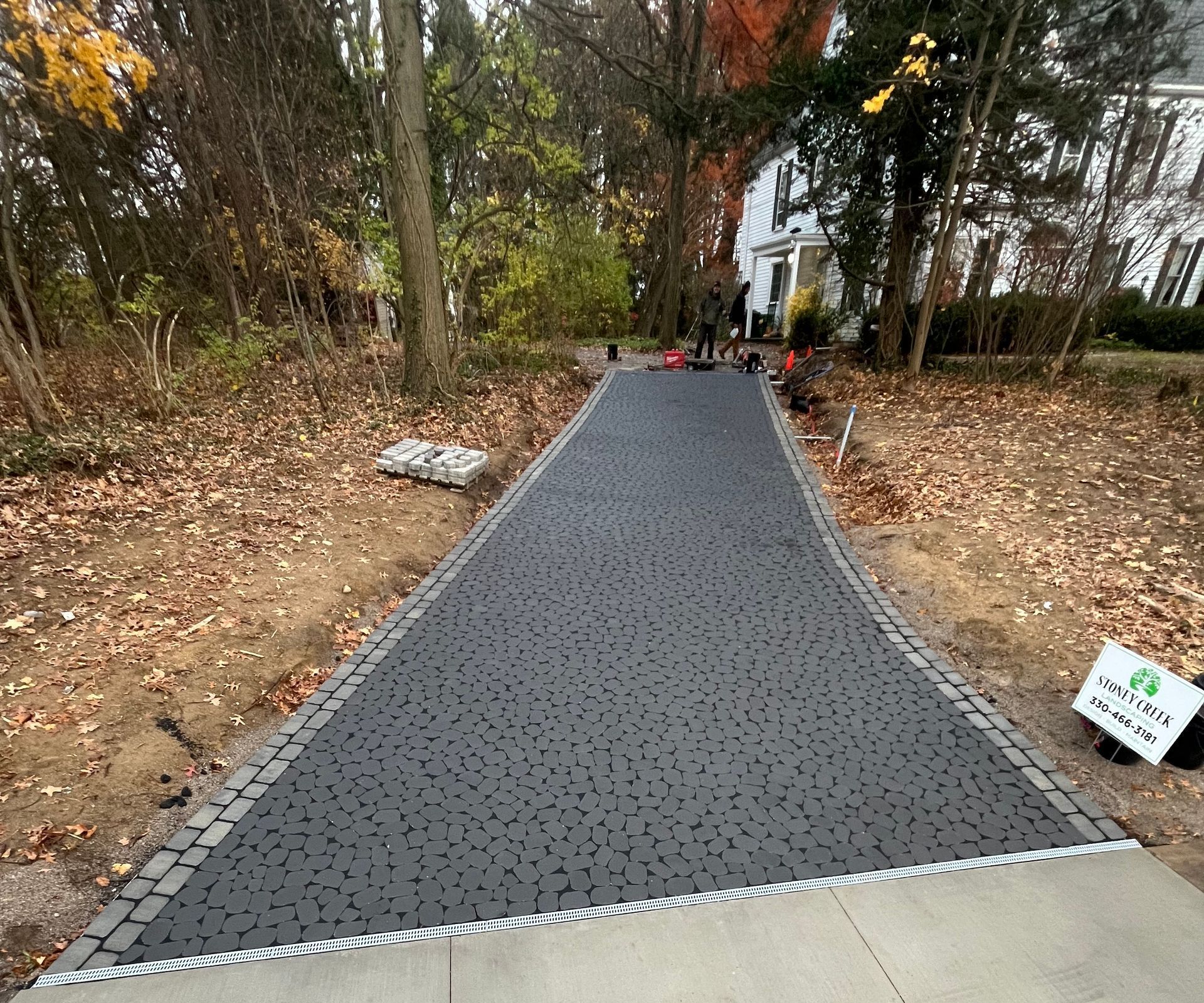 Asphalt driveway with gray stone border, leading to a house. Fall foliage surrounds.