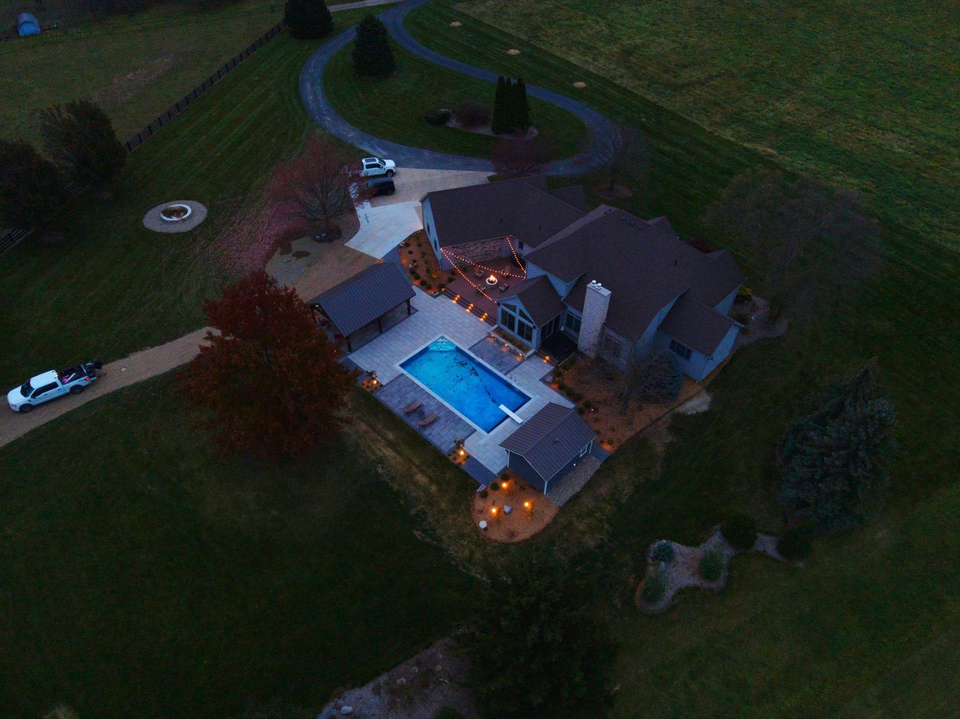 Aerial view of a house with a pool and circular driveway, lit by outdoor lights in a rural setting.