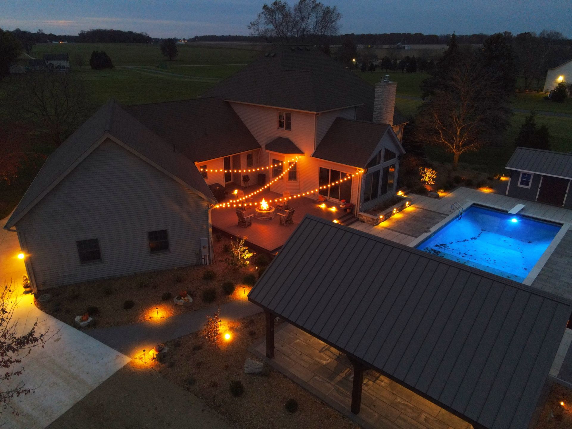 Nighttime aerial view of a house with a lit pool, patio, and string lights, surrounded by landscaping.