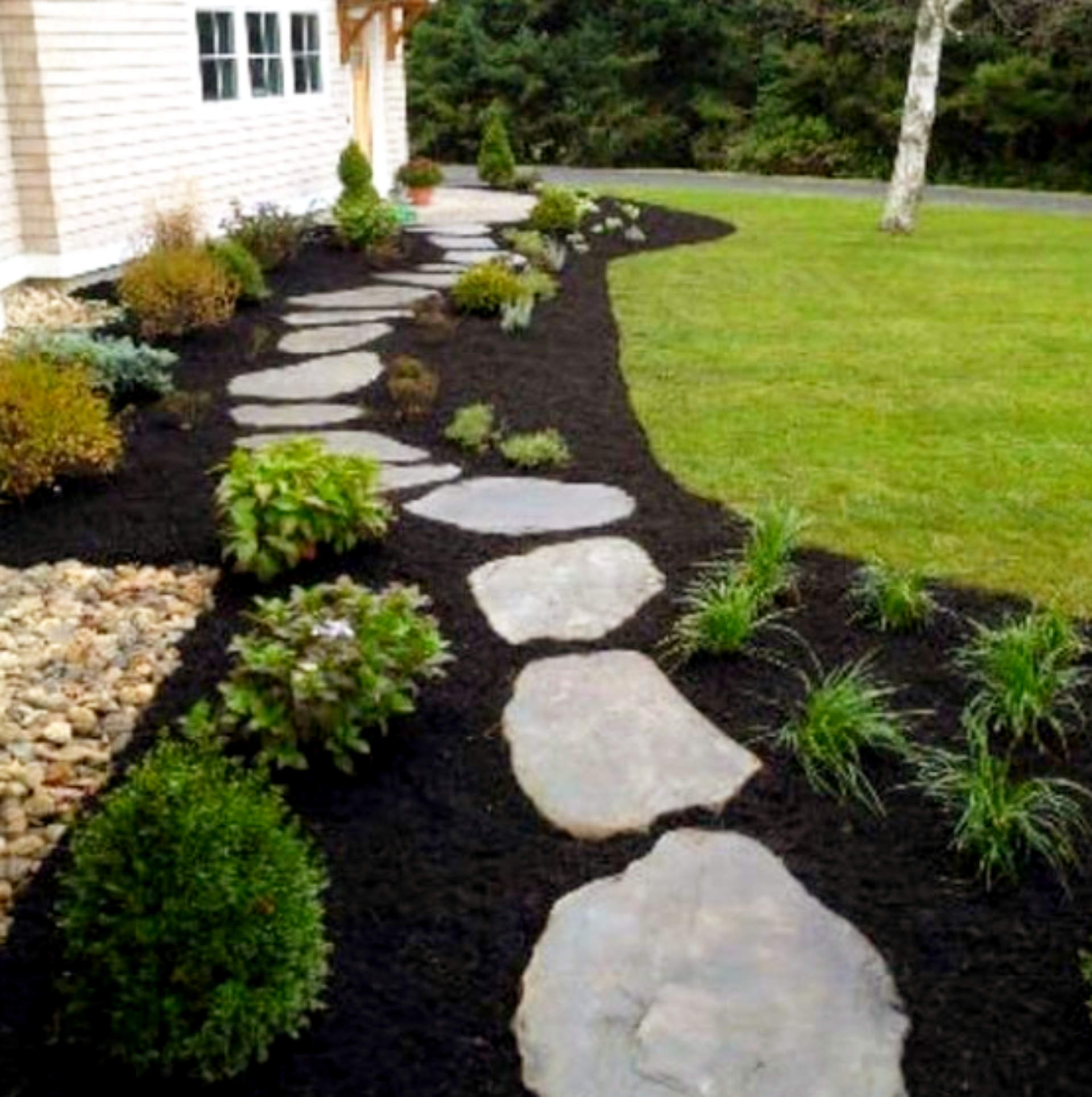 Stone path winds through garden with dark mulch and various plants beside a house and grassy lawn.