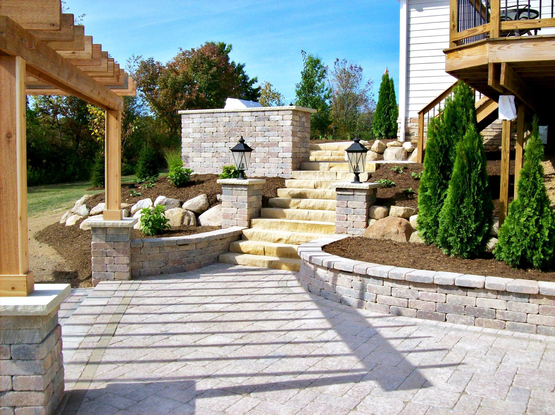 Stone patio with steps leading to a raised structure; wooden pergola on the left.