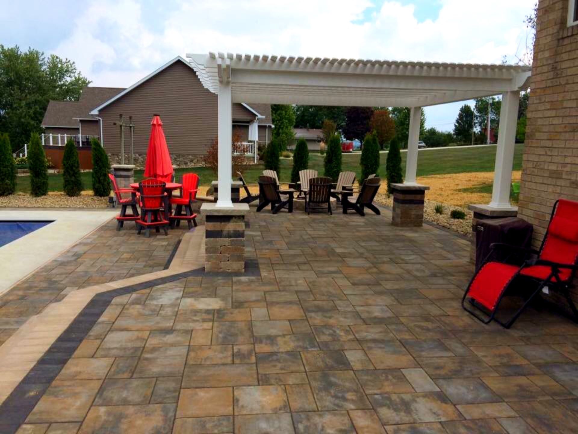 Patio with pergola, seating, and red umbrella by the pool. Brick home in background.