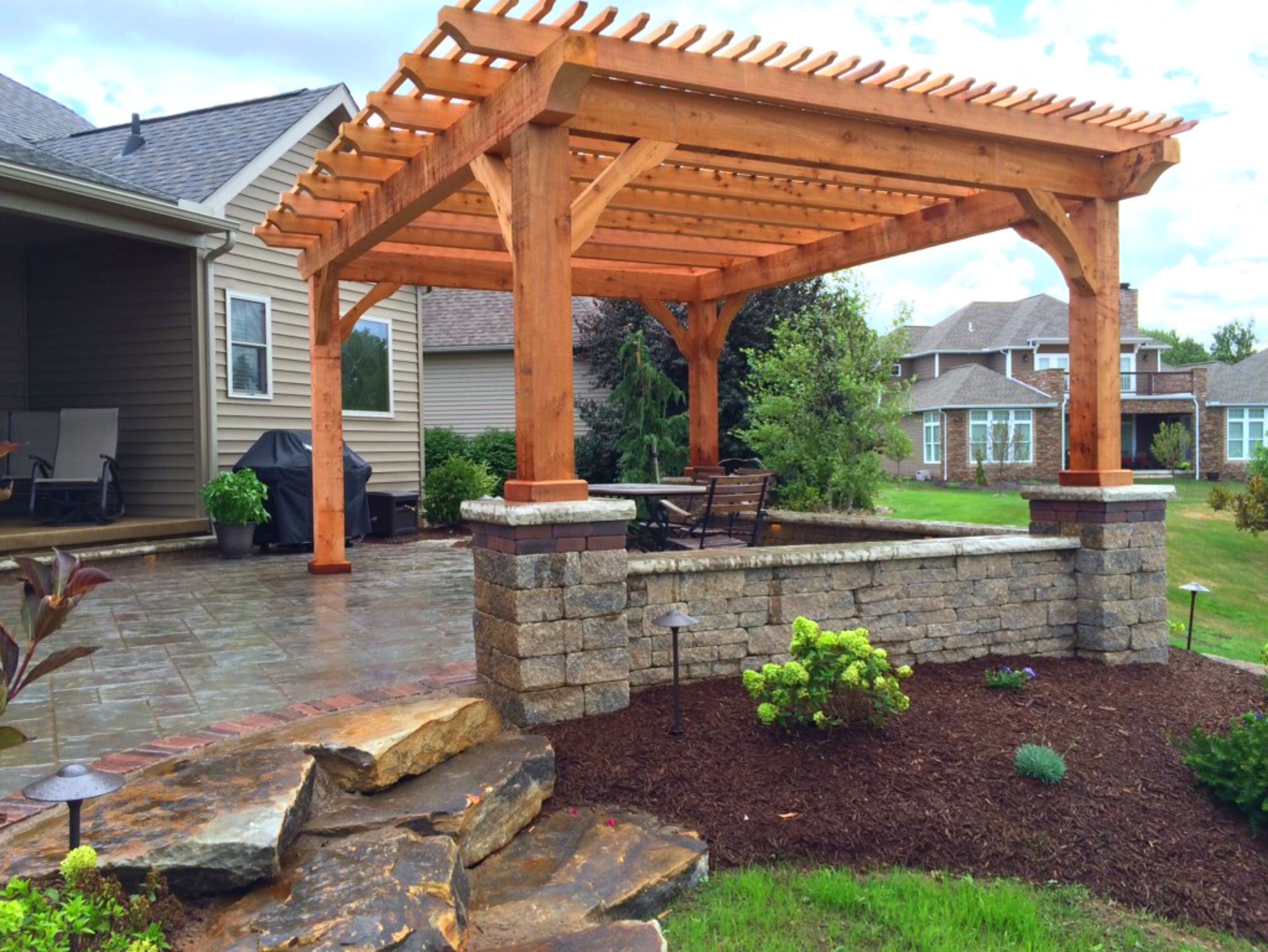 Wooden pergola over a stone patio with a low wall, backyard setting.