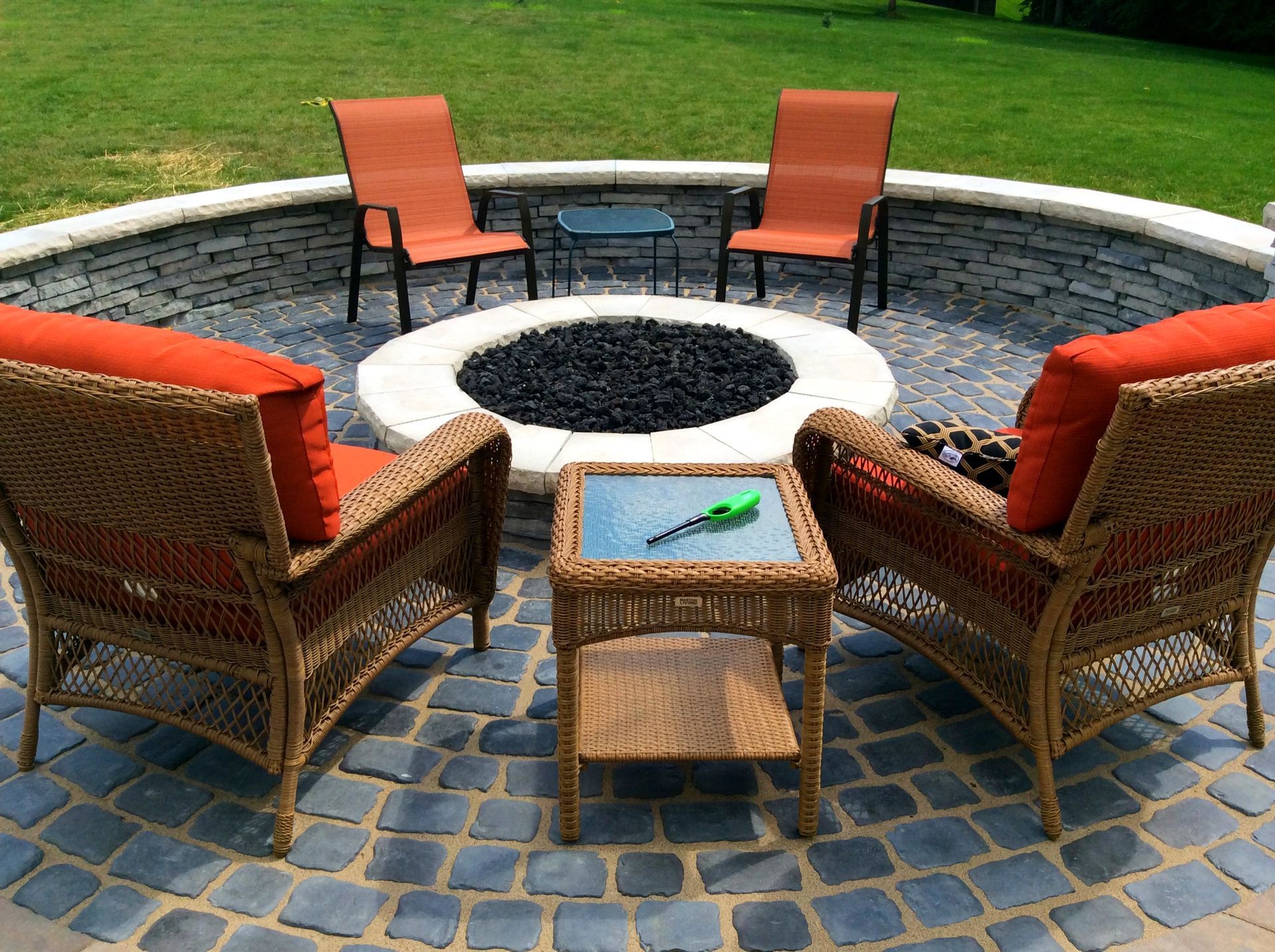 Fire pit area with wicker chairs and small tables on a circular stone patio. Orange cushions.
