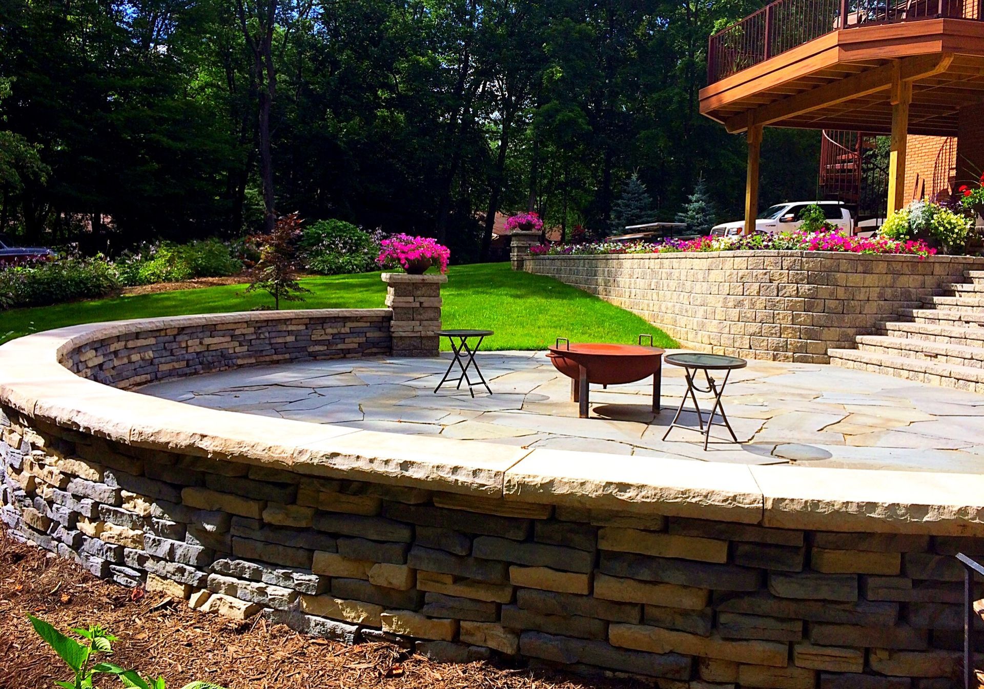 Curved patio with stone walls, grass, and a deck in the background, with flowers and seating.