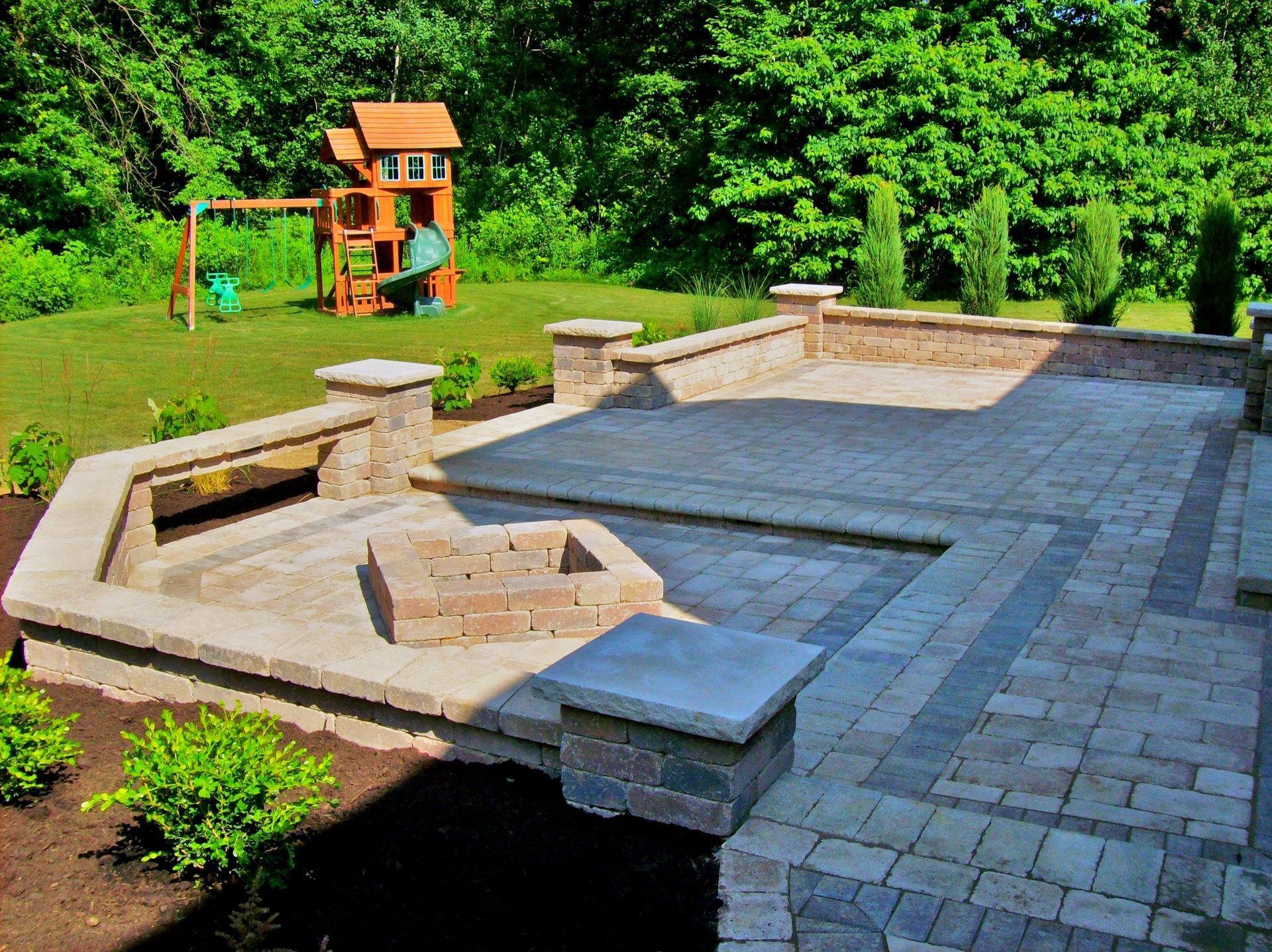 Outdoor patio with built-in fire pit and retaining walls, leading to a lawn with a playset in the background.
