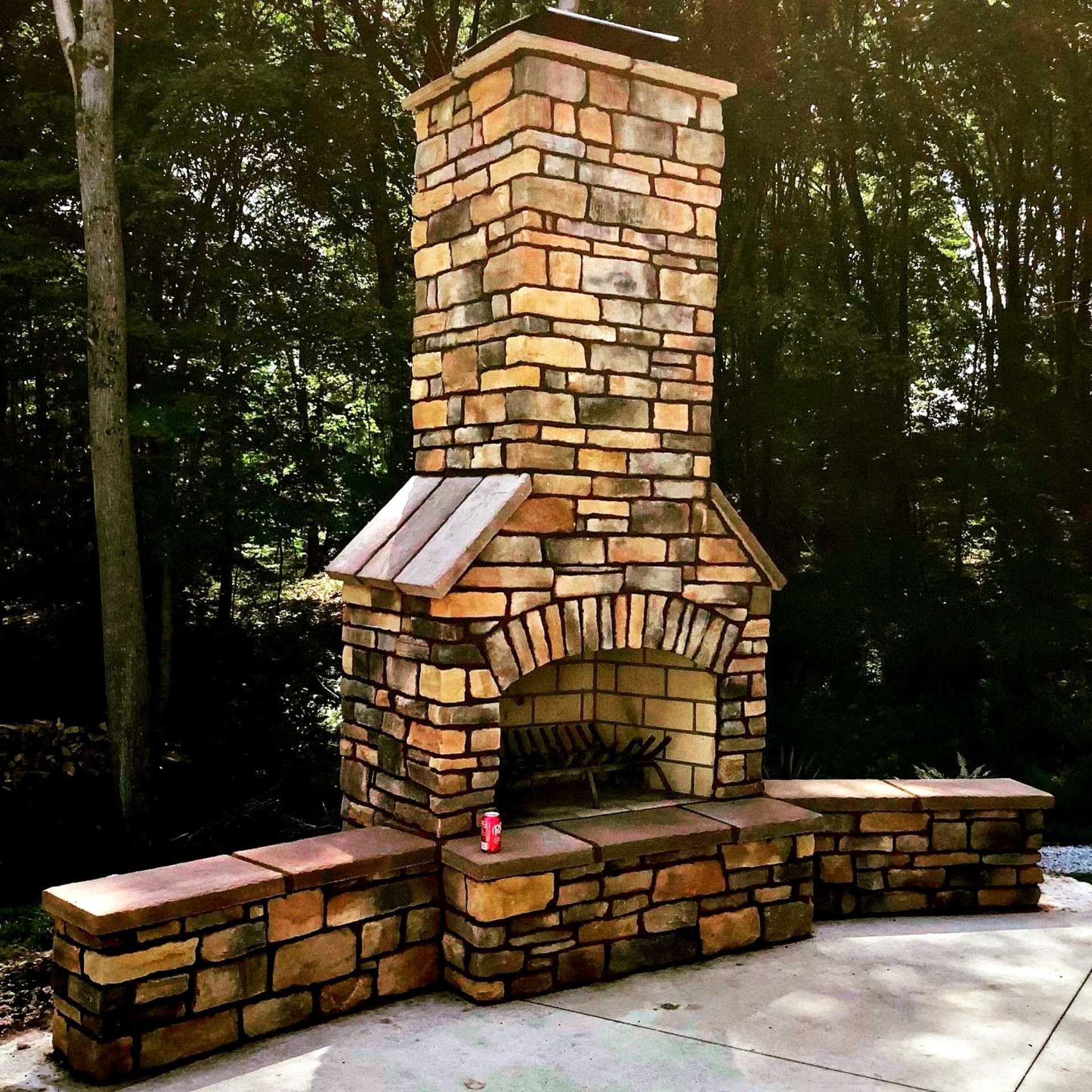 Stone outdoor fireplace with chimney, built-in benches, set on a patio, surrounded by trees.