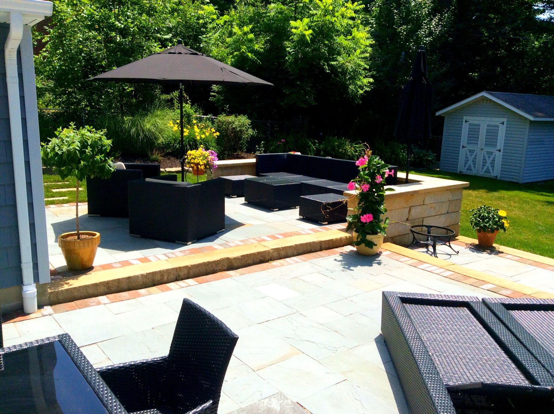 Backyard patio with black wicker furniture, umbrella, and potted plants on a sunny day.