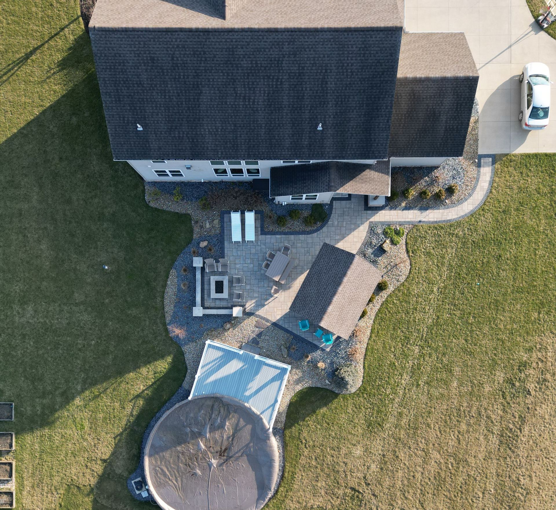 Overhead view of a house with a patio, fire pit, and pool cover on a grassy lot with a car parked in the driveway.