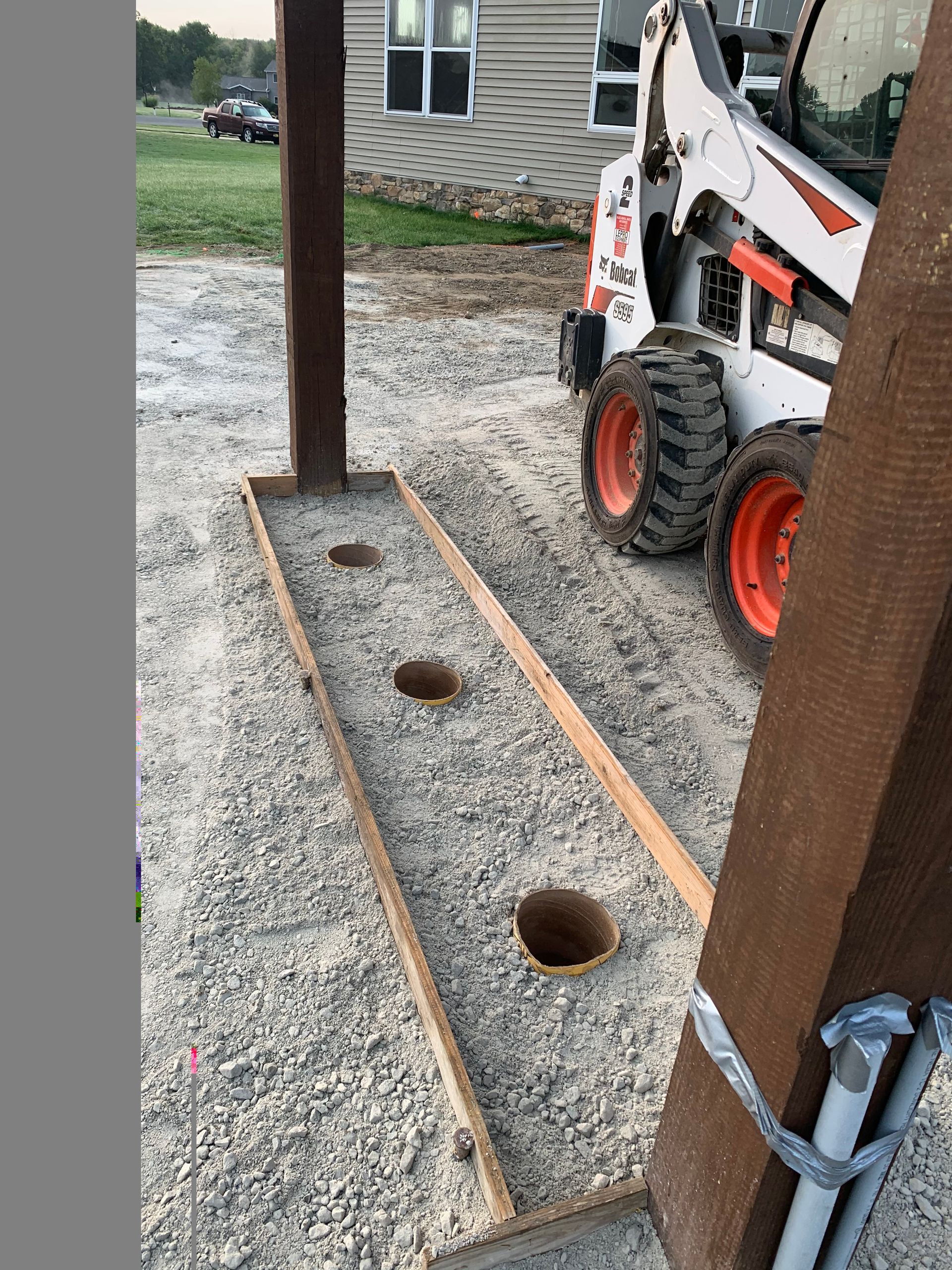 Construction site: Concrete form with three holes, gravel ground, Bobcat, and building.