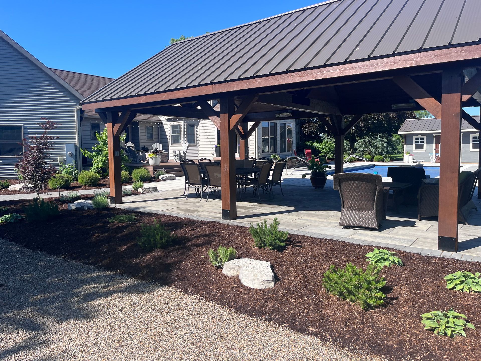 Patio with pergola over dining set, pool in background, brown mulch and landscaping.