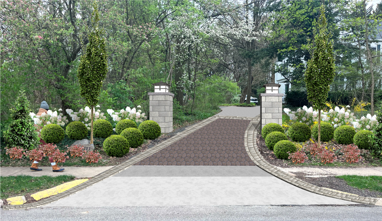 Driveway entrance with landscaping, featuring stone columns and small trees.