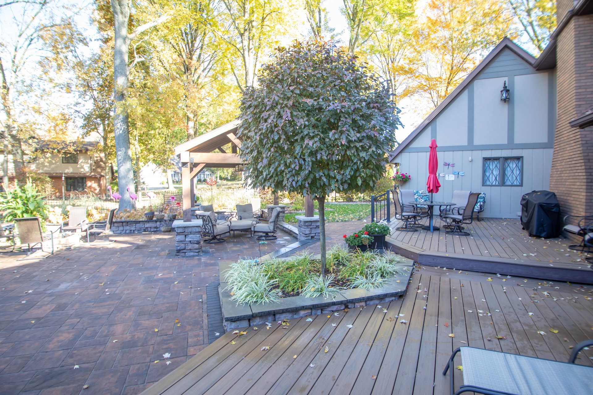 Backyard patio with deck, seating, and a tree. Brown deck leads to stone patio with gazebo and house.