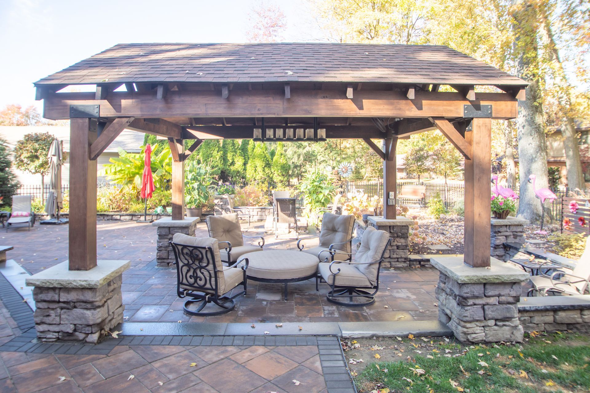 Patio with pergola, stone columns, seating area. Brown wood, stone pavers, lush greenery.