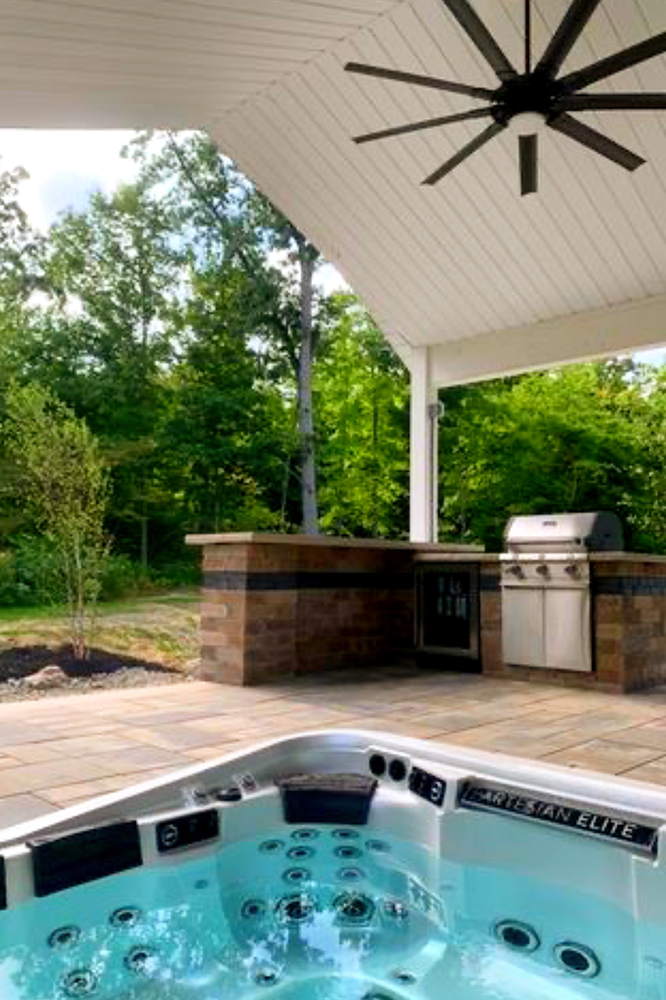 Outdoor kitchen with hot tub. Patio under a white-ceilinged overhang with a built-in grill and a dark ceiling fan.