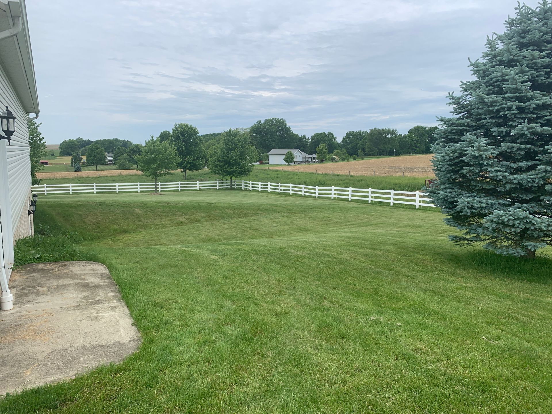 Lush green yard with white fence and trees leading to a cloudy countryside view.