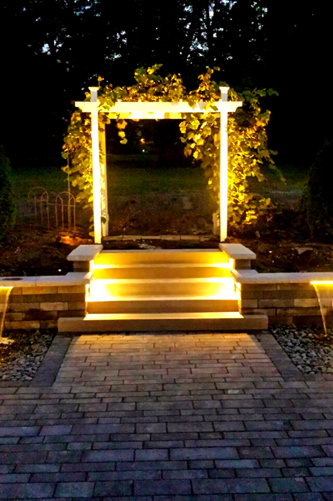 Lit white pergola with vine and steps, warm lighting on a brick pathway.