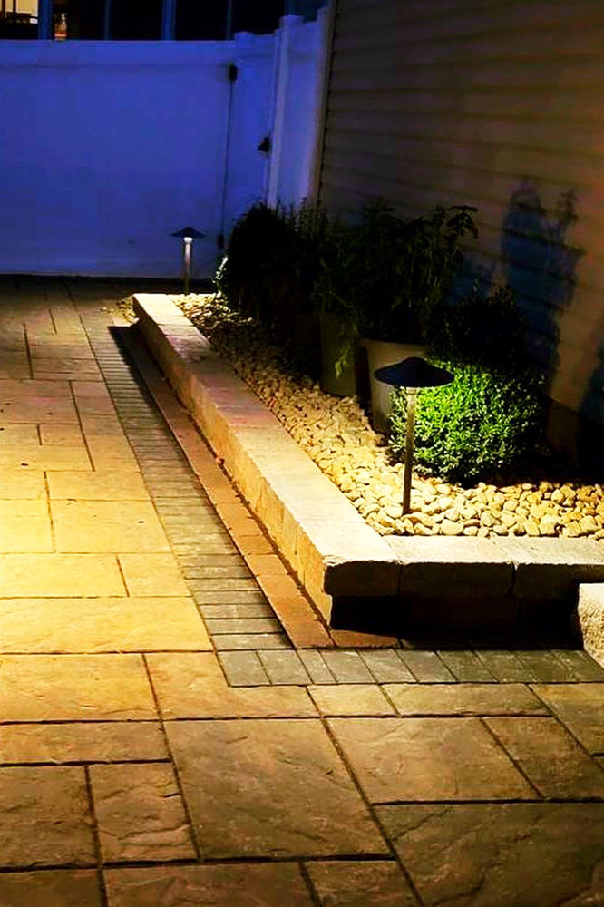 Lit outdoor patio with stone pavers, brick border, and illuminated landscaping at night.
