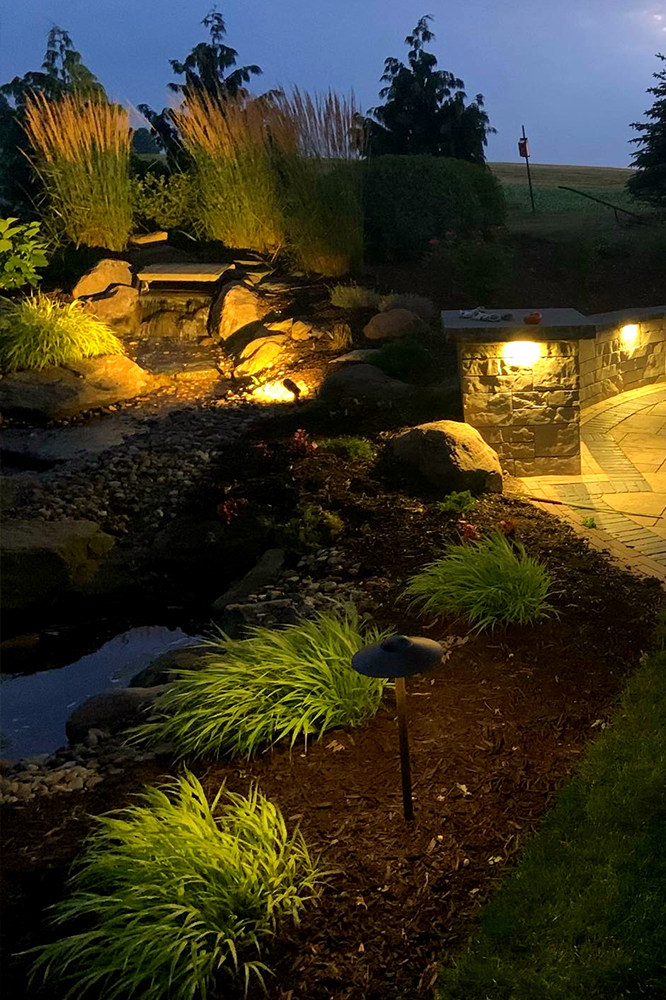 Nighttime landscape with illuminated plants, rocks, and a water feature.