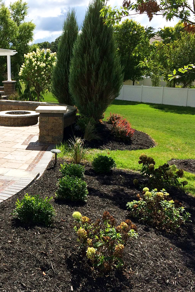 Landscaped backyard with trees and flowerbeds, brick patio, fire pit, and white fence.