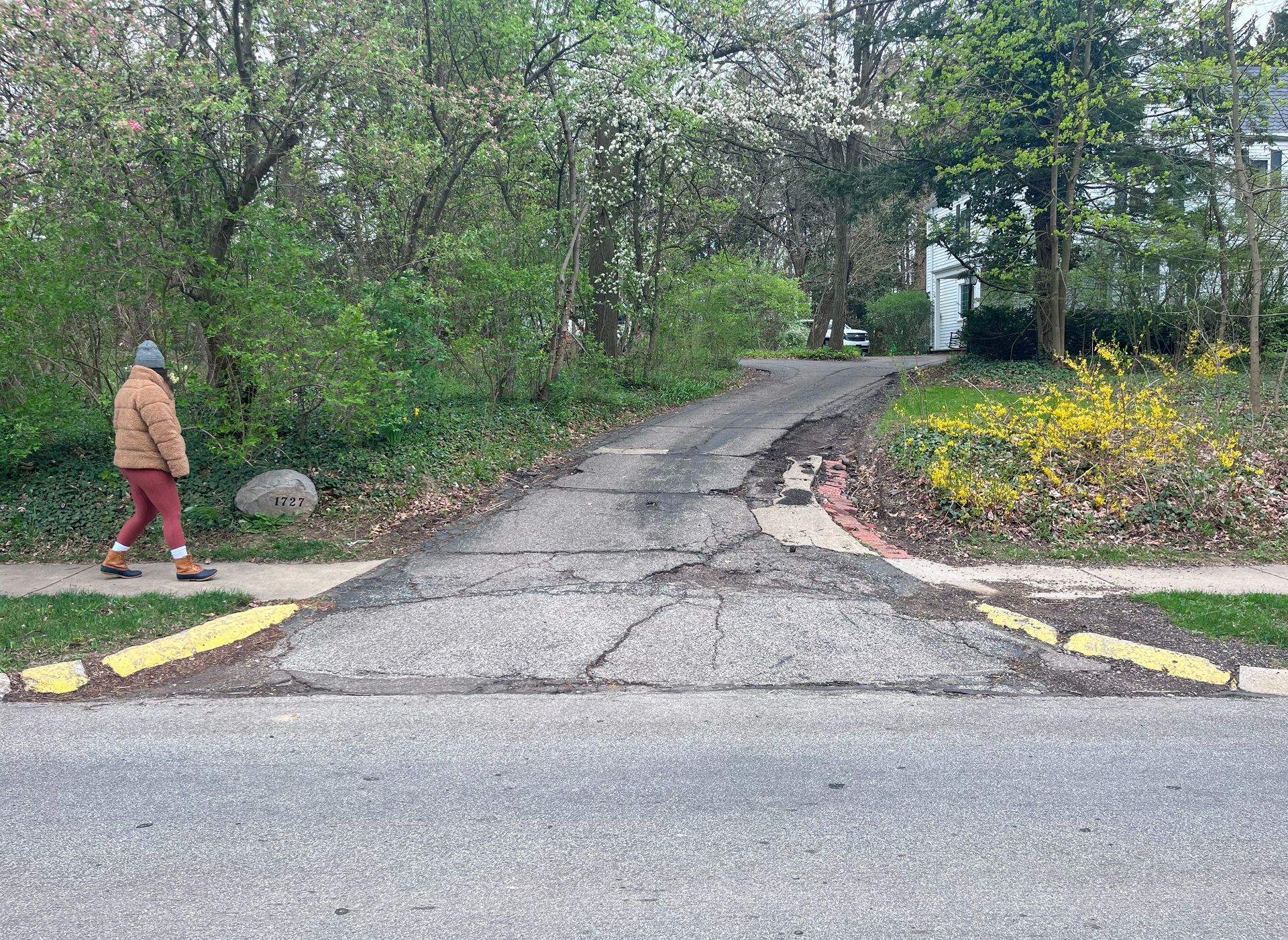 Cracked driveway leads to trees and a house; person walks on the sidewalk.