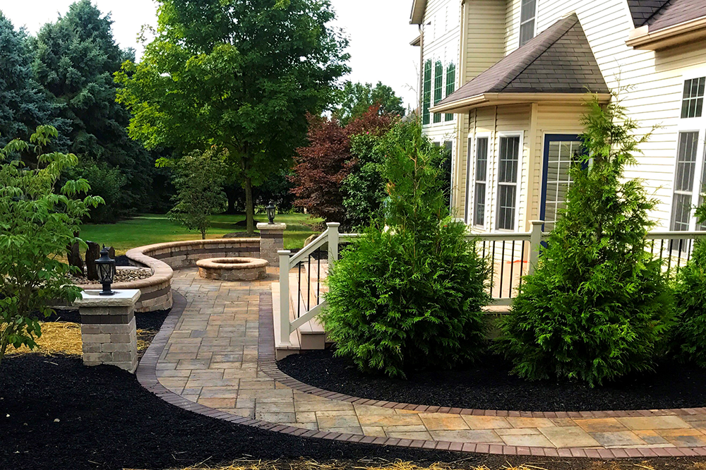Stone walkway leads to backyard patio with fire pit, bordered by evergreens and the side of a house.