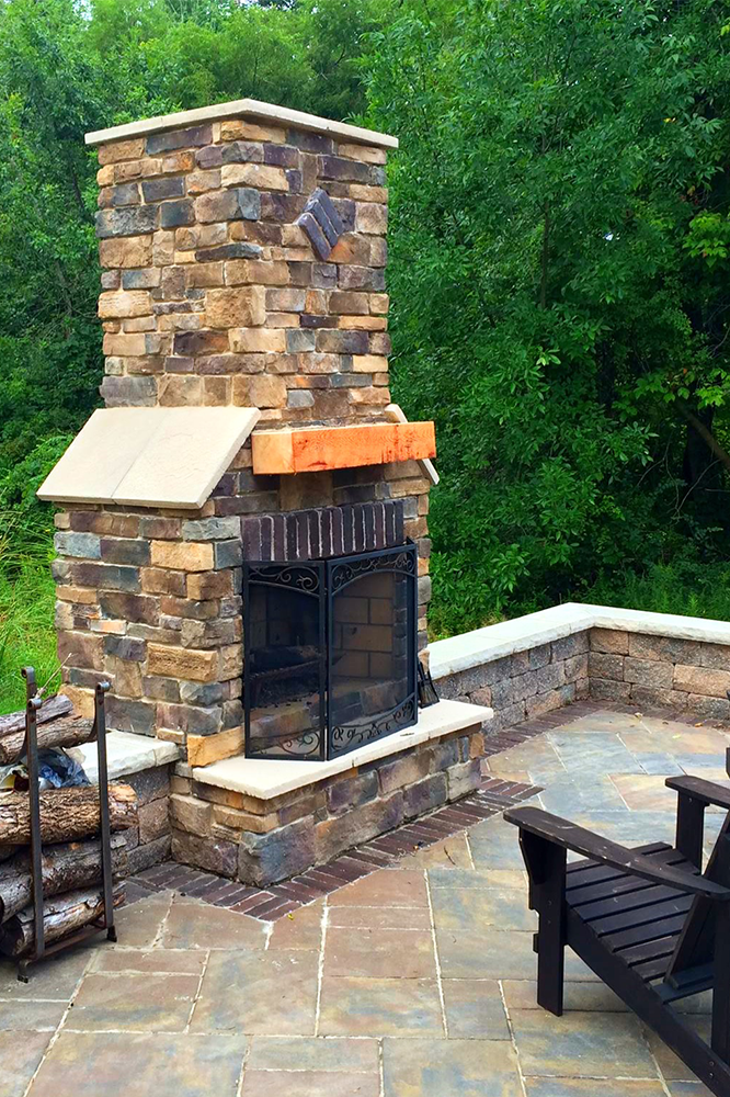 Stone outdoor fireplace on a patio with wood stack and green trees in background.