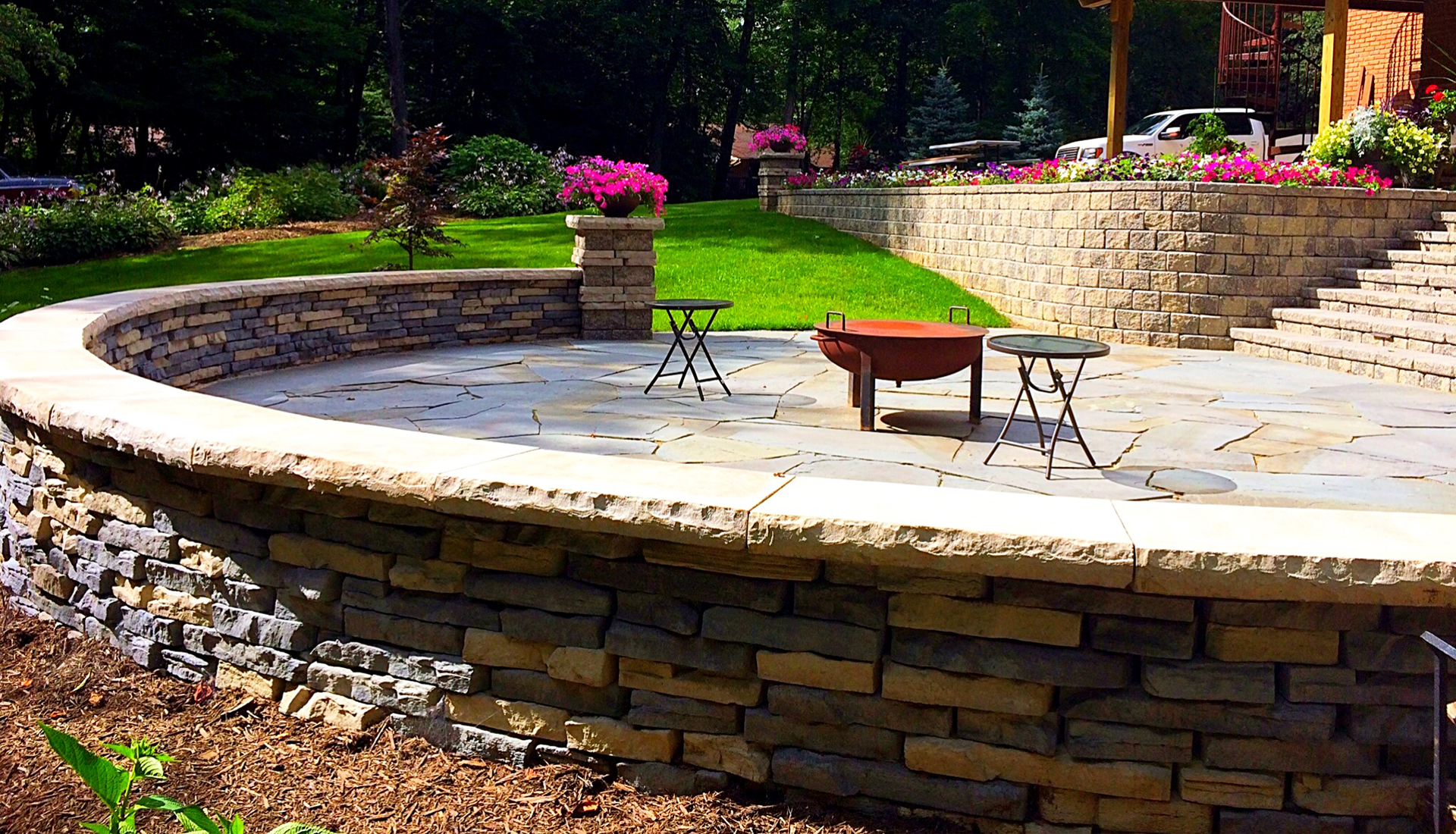 Curved stone patio with fire pit, small tables, and surrounding retaining walls, lush landscaping.